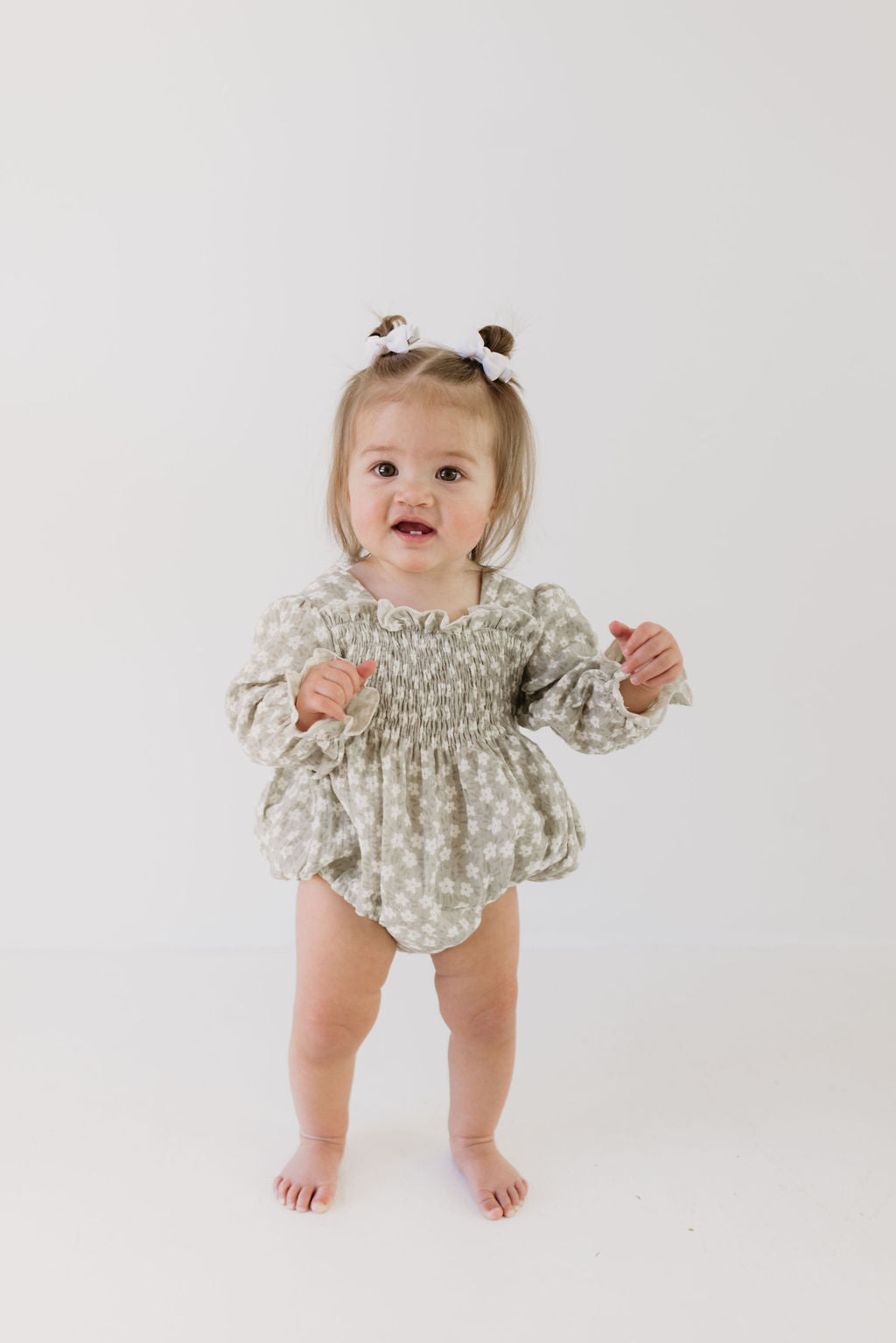 A smiling toddler in a French Gray Floral Ruffle Romper by forever french baby stands against a plain white background. Her hair is styled in two buns with white bows, perfectly complementing her infant-sized outfit.