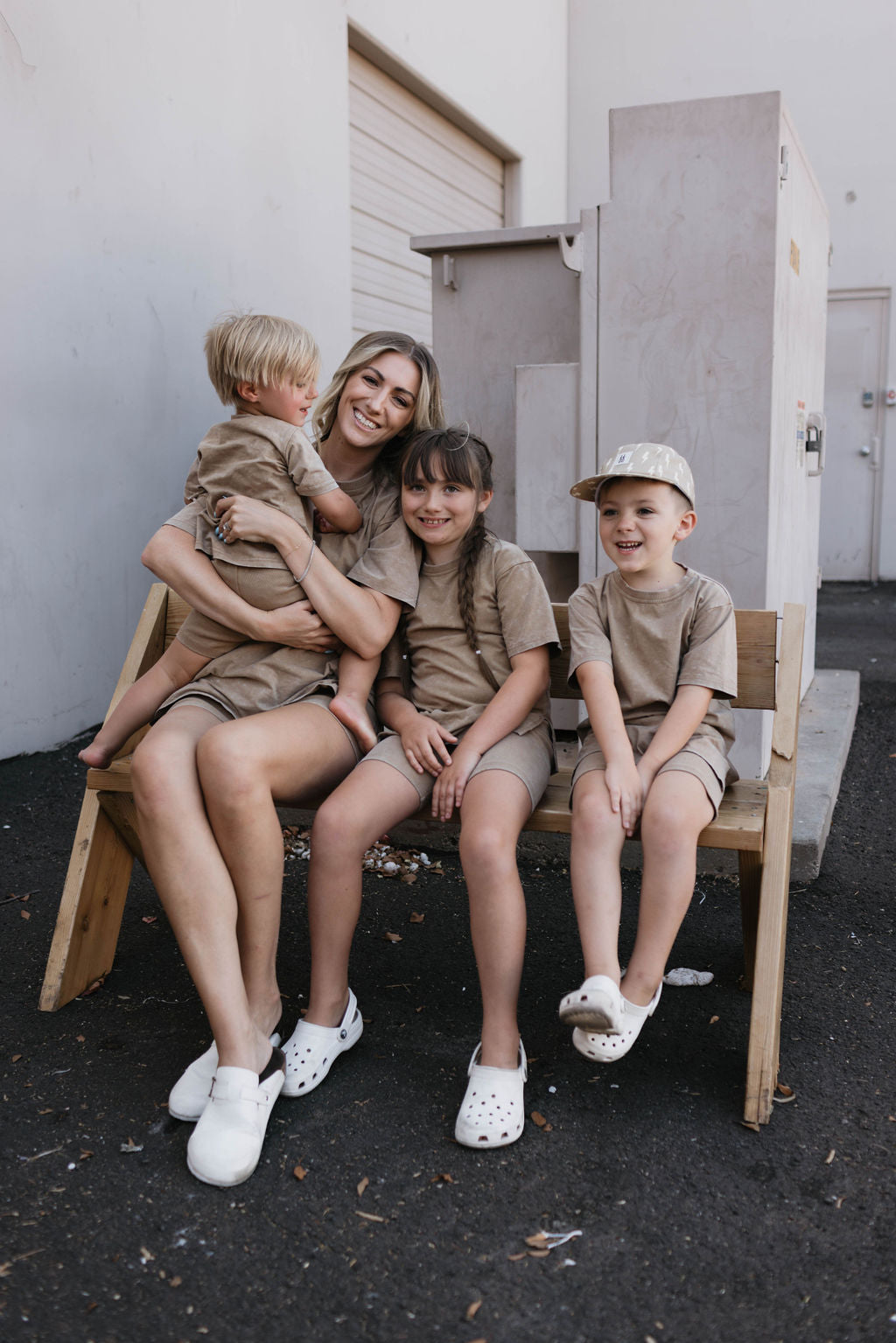 A woman sits on a wooden bench outdoors, smiling and holding a small child dressed in an outfit from the forever french baby collection. Two other children sit beside her, all wearing matching beige outfits. One child complements their attire with a Vintage Washed Espresso cap and white Crocs. They all appear relaxed and happy in their coordinated look featuring items from the Women's Short Set | Vintage Washed Espresso by forever french baby.