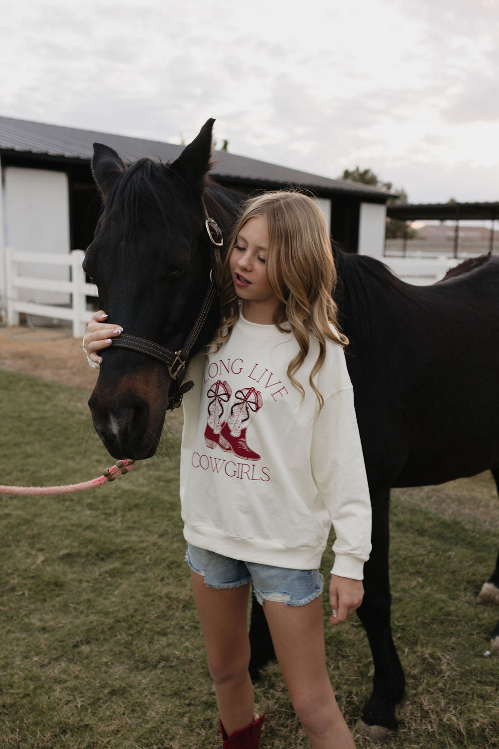 A girl with long hair, dressed in a cozy "Kids Crewneck | Long Live Cowgirls" by lolo webb and denim shorts, stands next to a black horse on a grassy field. She gently holds the horse's face. In the background, there's a white fence and a stable. For those who appreciate cotton elastane comfort, this sweatshirt is available in an adult version as well.