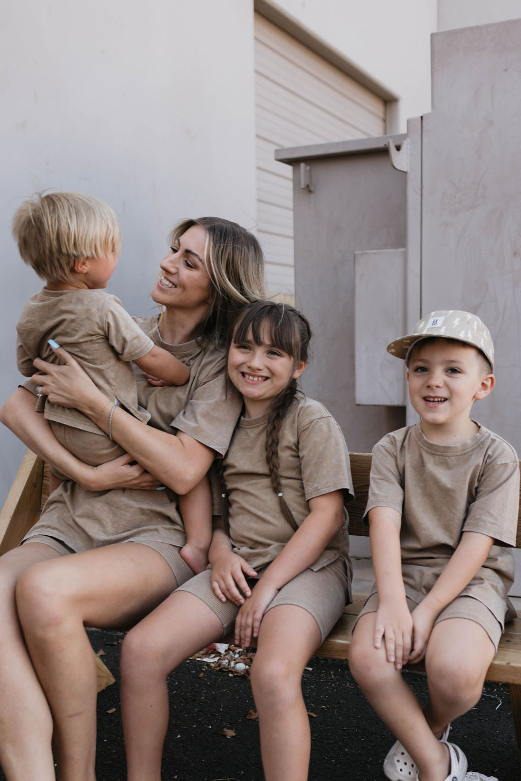 A woman sits on a wooden bench with three young children, all wearing matching beige outfits from forever french baby. The woman is dressed in the Women's Short Set in Vintage Washed Espresso and is holding the youngest child, who has blond hair. The two older children sitting next to her are smiling; one has braided hair and the other wears a cap.