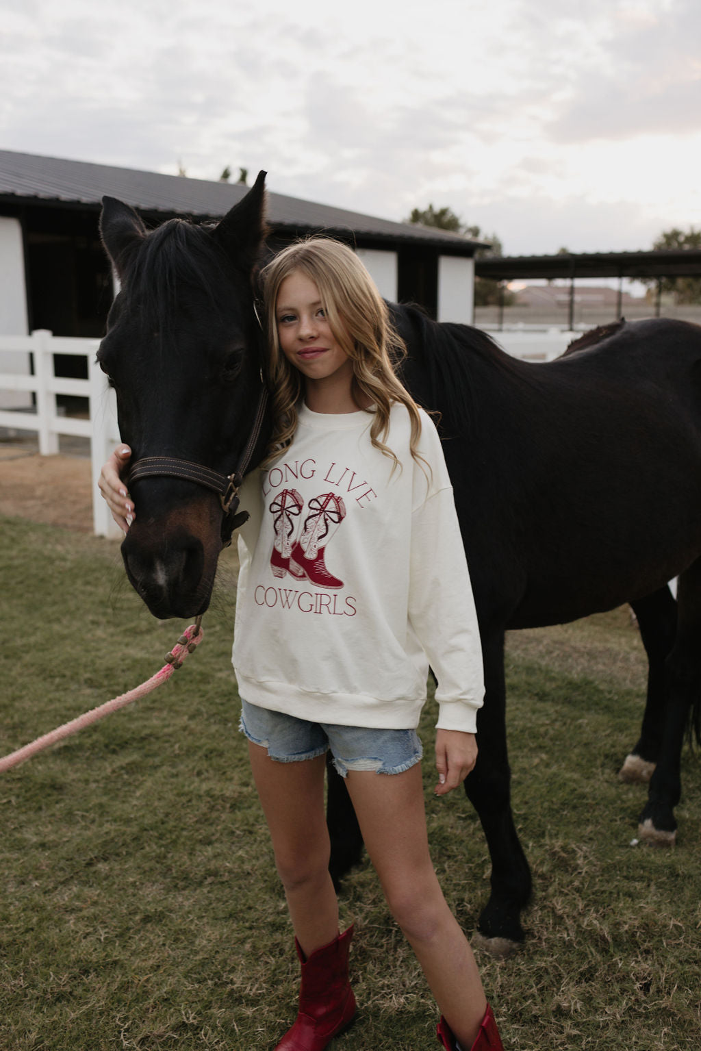 A girl stands beside a black horse, tenderly holding its head. She's wearing a lolo webb Kids Crewneck sweater with "Long Live Cowgirls" along with denim shorts, and finishes the look with red boots. They're in a grassy area, framed by a stable in the background, capturing an ideal holiday moment.