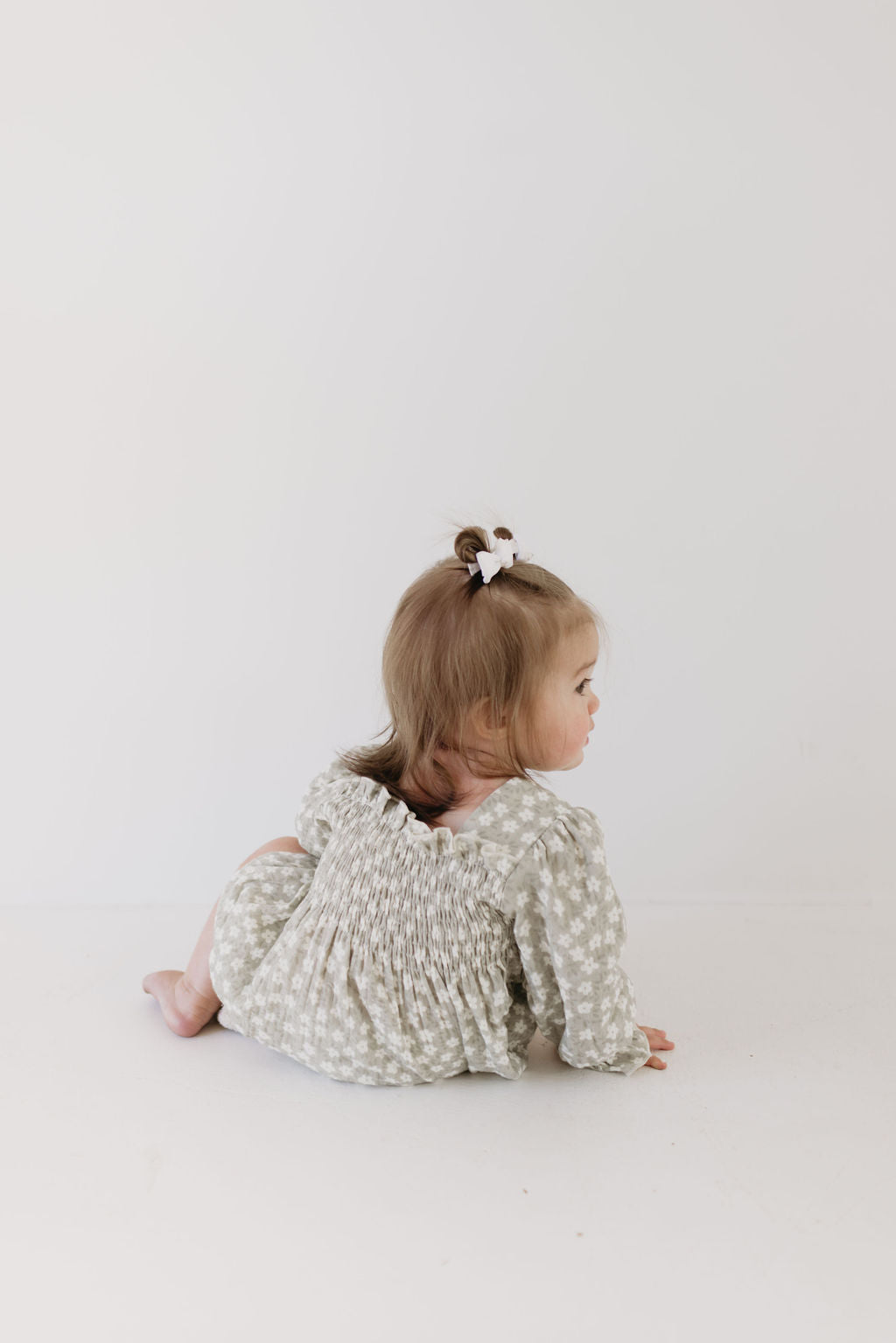 A toddler with light hair sits on a white surface, wearing the Ruffle Romper in French Gray Floral by forever french baby, complete with a bow in her hair. She gazes over her shoulder to the right side of the image.