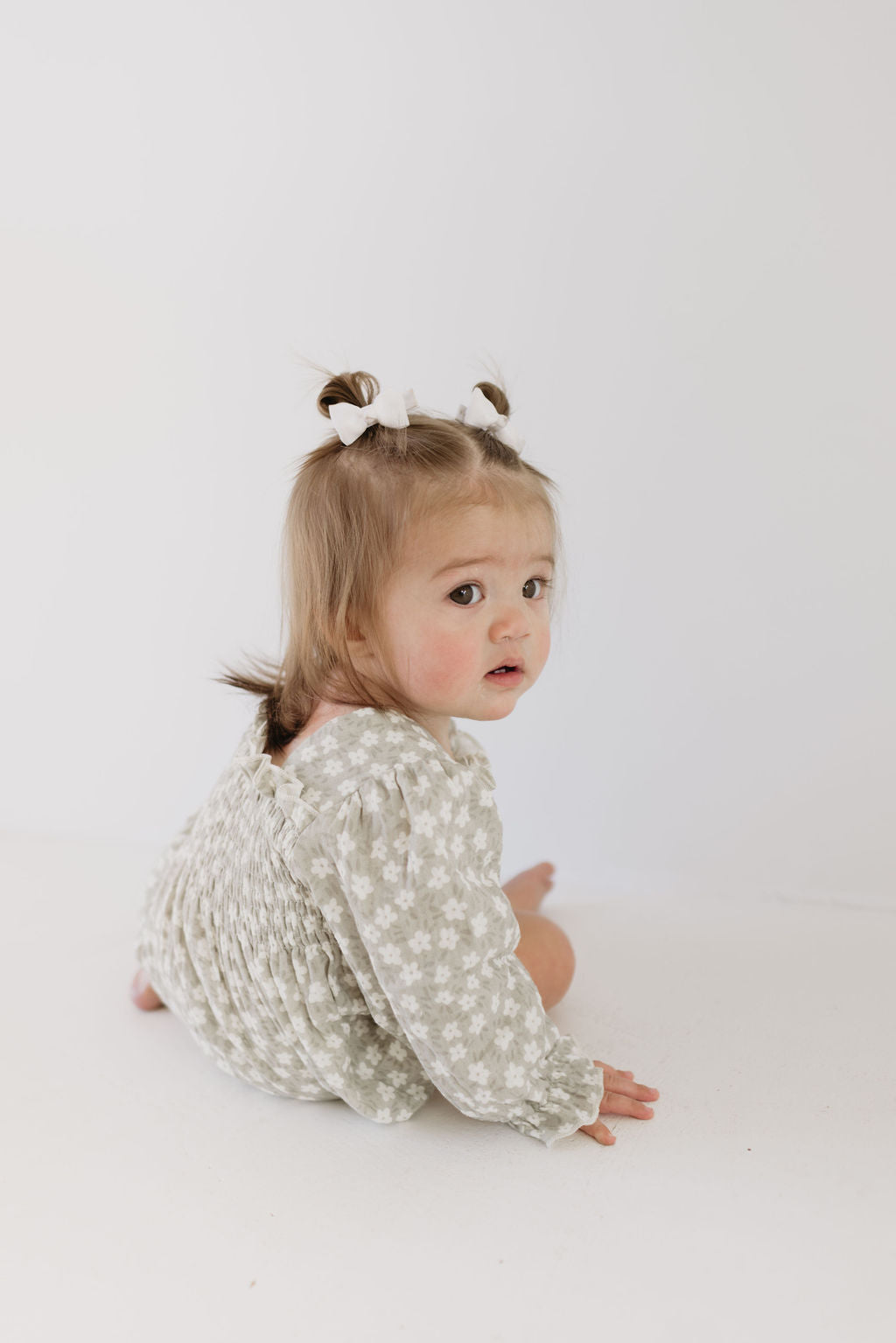 A toddler with pigtails and white bows wears a forever french baby Ruffle Romper in French Gray Floral, sitting on the ground and looking over their shoulder against a light background.