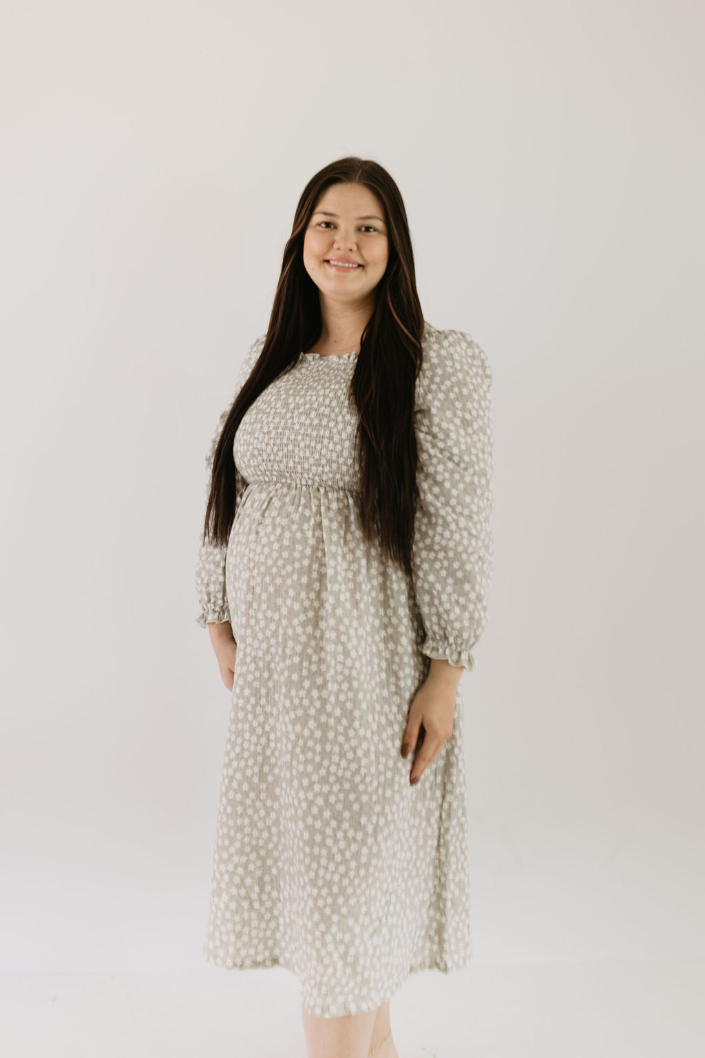 A pregnant woman with long dark hair smiles while wearing a "forever french baby" Adult Dress in French Gray Floral. She stands against a plain white background, exuding soft elegance that complements matching rompers perfectly.