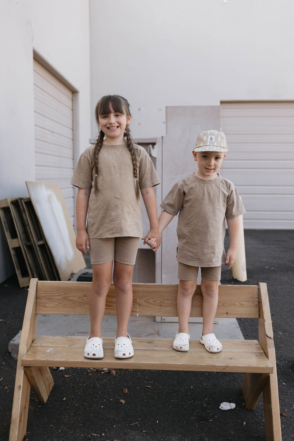 Two children stand on a wooden bench holding hands, wearing matching Children's Short Sets in Vintage Washed Espresso from the forever french baby collection, paired with white Crocs. The child on the left has long braided hair, while the one on the right wears a baseball cap. They are outside in front of a building with a closed garage door.