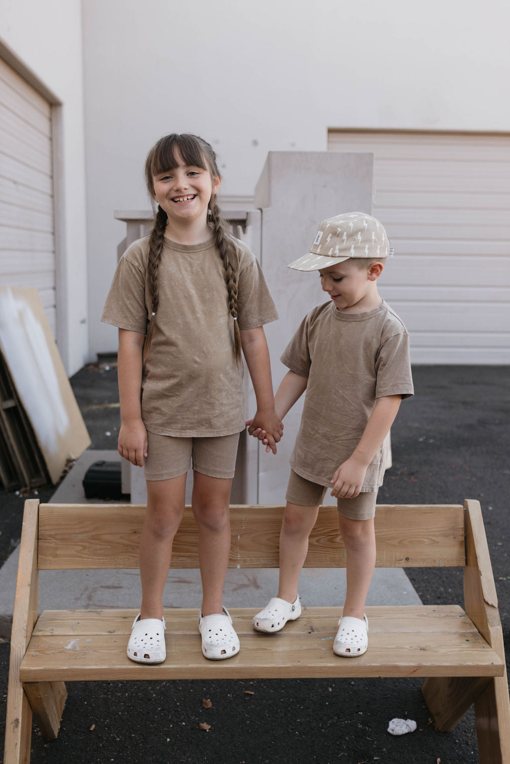 Two children are standing on a wooden bench holding hands and smiling. The girl on the left has long braided hair and is wearing a Forever French Baby beige children's summer outfit with white shoes. The boy on the right, in a matching Forever French Baby Children's Short Set in Vintage Washed Espresso, wears a cap and white shoes, looking down.