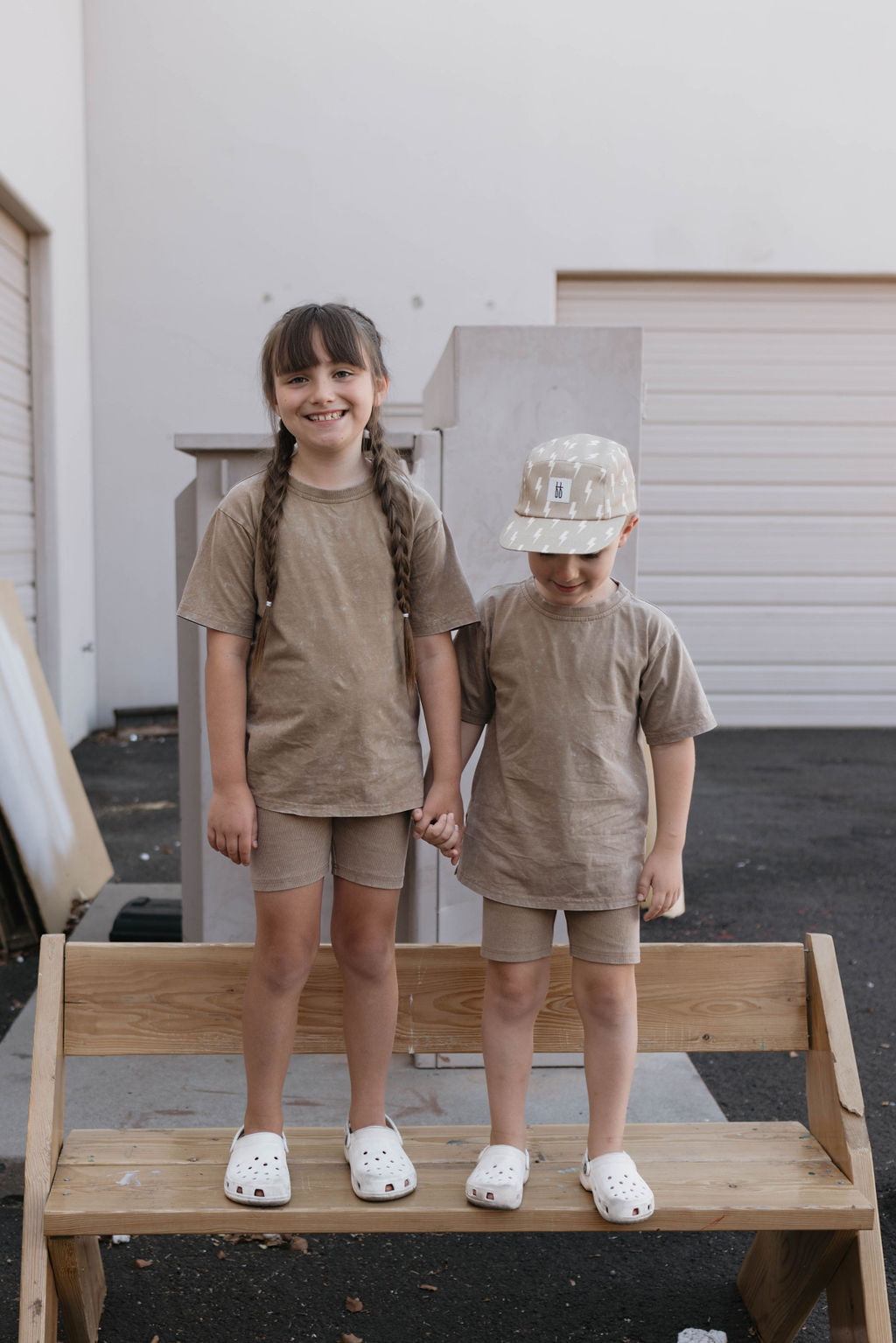 Two children are standing and holding hands on a bench outdoors, both wearing matching Vintage Washed Espresso short sets from forever french baby, paired with white shoes. The older child with long braided hair and the younger one in a beige helmet stand before a charming white building.