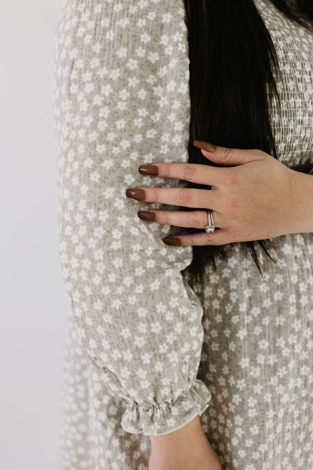 Wearing a forever french baby Adult Dress in French Gray Floral with textured fabric, a person gently holds their arm. Their brown hair cascades elegantly as they showcase a silver ring on their left finger.