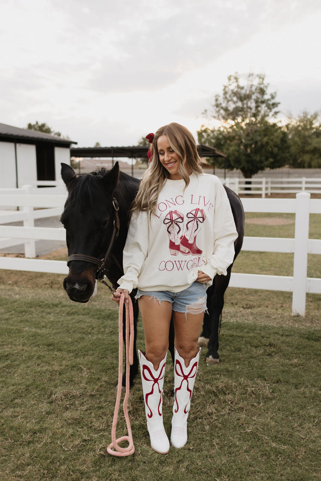 A woman in denim shorts and white boots with red designs stands next to a black horse. She is wearing the "Long Live Cowgirls" Adult Crewneck by lolo webb, made of soft cotton. They are in a grassy, fenced area with trees and buildings visible in the background.