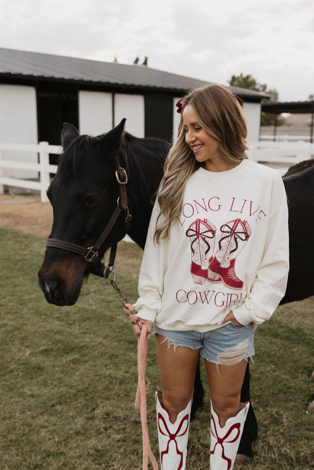 A woman in an Adult Crewneck "Long Live Cowgirls" sweater by lolo webb and cowboy boots is smiling beside a black horse. She's standing on grass in front of a white fence and barn, with her long hair loose as she holds the horse's lead rope, perfectly coordinating with the children's version of her outfit.