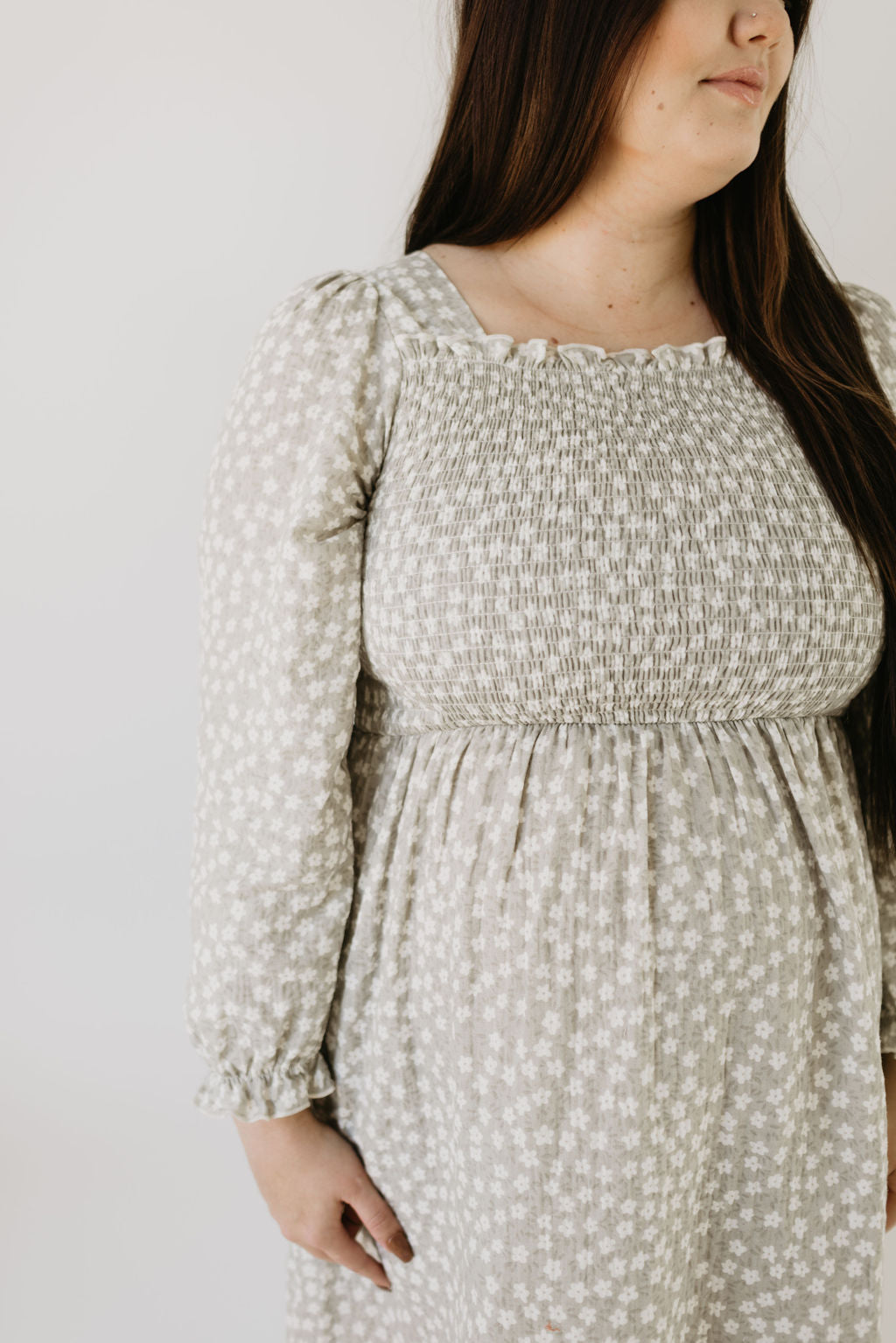 A woman models the forever french baby Adult Dress in French Gray Floral, showcasing its long sleeves and shirred bodice against a simple light backdrop, with her long dark hair elegantly draped over one shoulder.