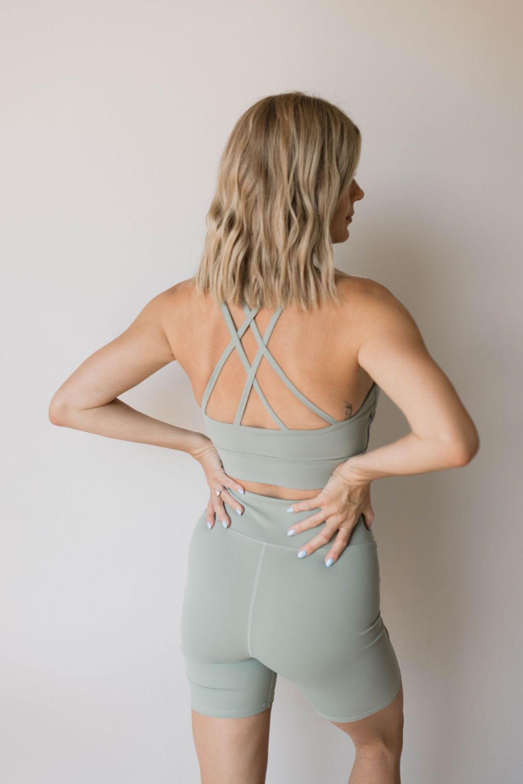 A person with shoulder-length blonde hair stands against a plain backdrop, facing away. They are wearing the Women's Workout Set in green by forever french baby, which includes a light green sports bra with crisscross straps and matching high-waisted biker shorts. Their hands are placed on their lower back.