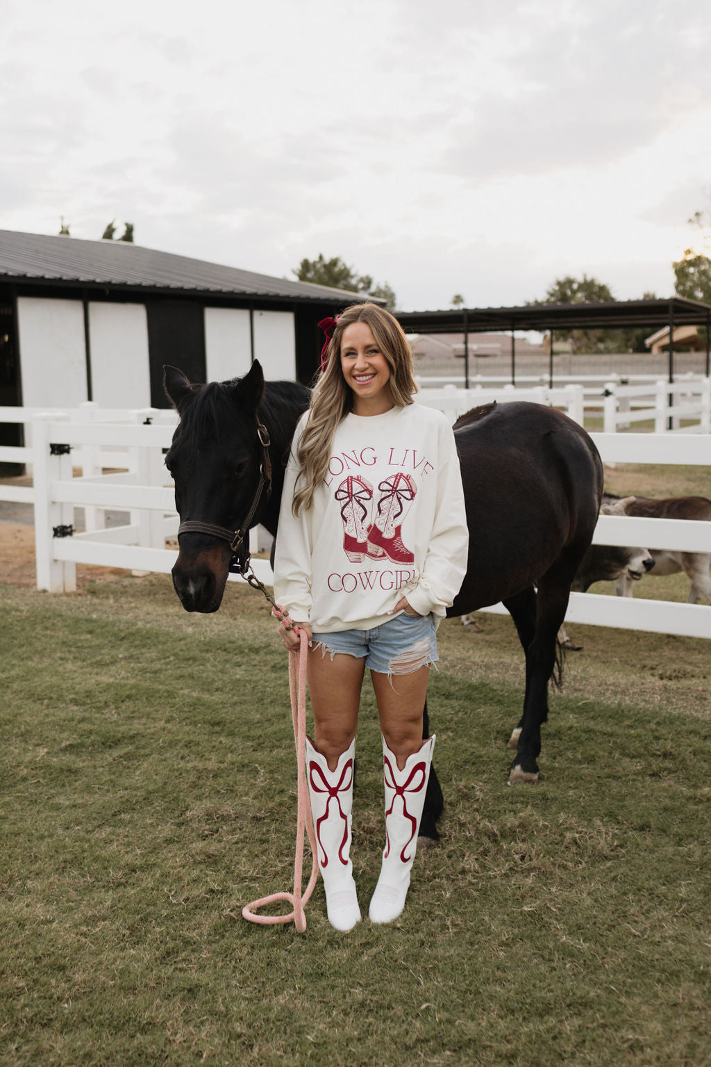 A woman is in a grassy area beside a black horse, dressed in the "Adult Crewneck | Long Live Cowgirls" by lolo webb, paired with denim shorts and white cowboy boots adorned with red designs. In the background, a white fence and barn set the scene for this timeless style.