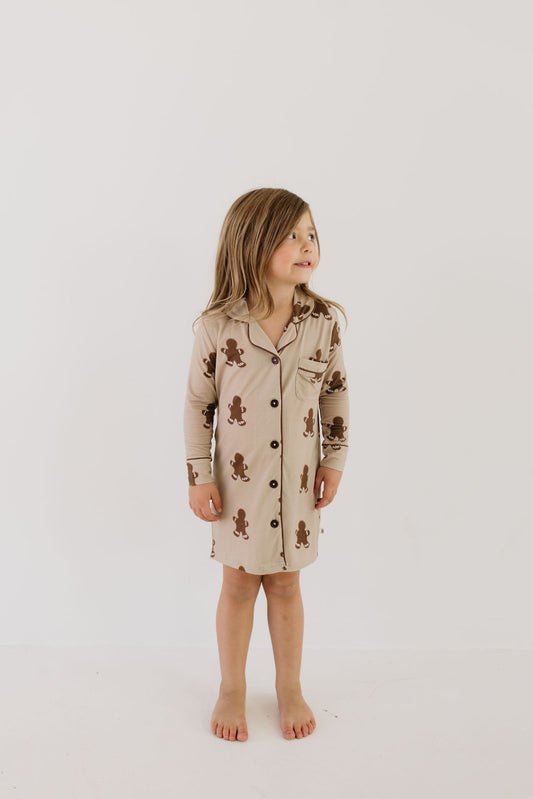 A young child with long hair stands barefoot against a plain white background, wearing the "Toddler Bamboo Sleeping Dress | Gingerbread" by forever french baby. This light brown sleeping dress, made from hypo-allergenic bamboo fabric, is decorated with a pattern of small dark brown gingerbread figures.