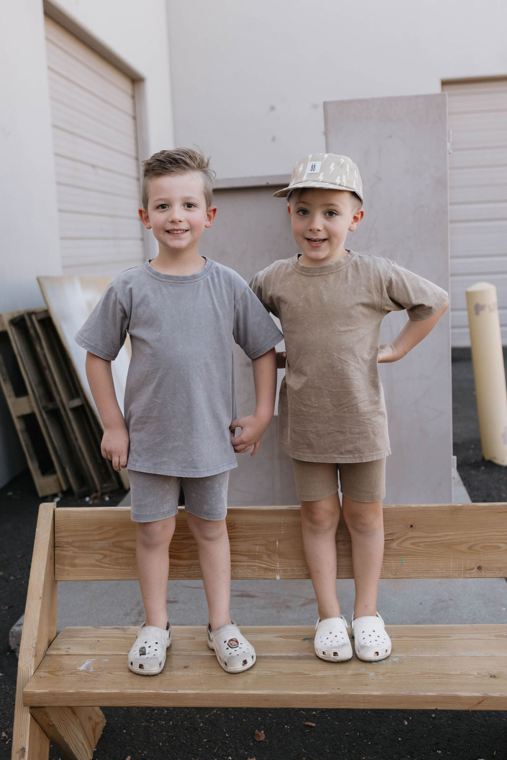 Two young children stand on a wooden bench, smiling at the camera in their neutral-toned outfits. One wears a cap, and both have on white Crocs. The child in the beige outfit sports a Children's Short Set in Vintage Washed Espresso from forever french baby. A light-colored building and wooden pallets serve as the backdrop.