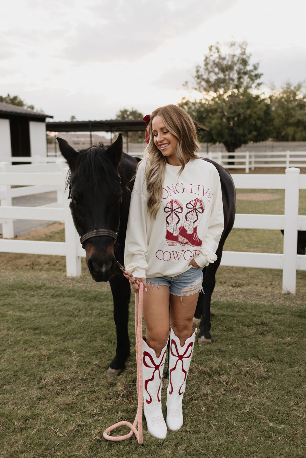 A person dressed in a "Long Live Cowgirls" Adult Crewneck by lolo webb, paired with denim shorts and white cowboy boots, stands next to a black horse on a grassy field. A matching children's version adds to the charming scene, framed by a white fence and trees in the background.