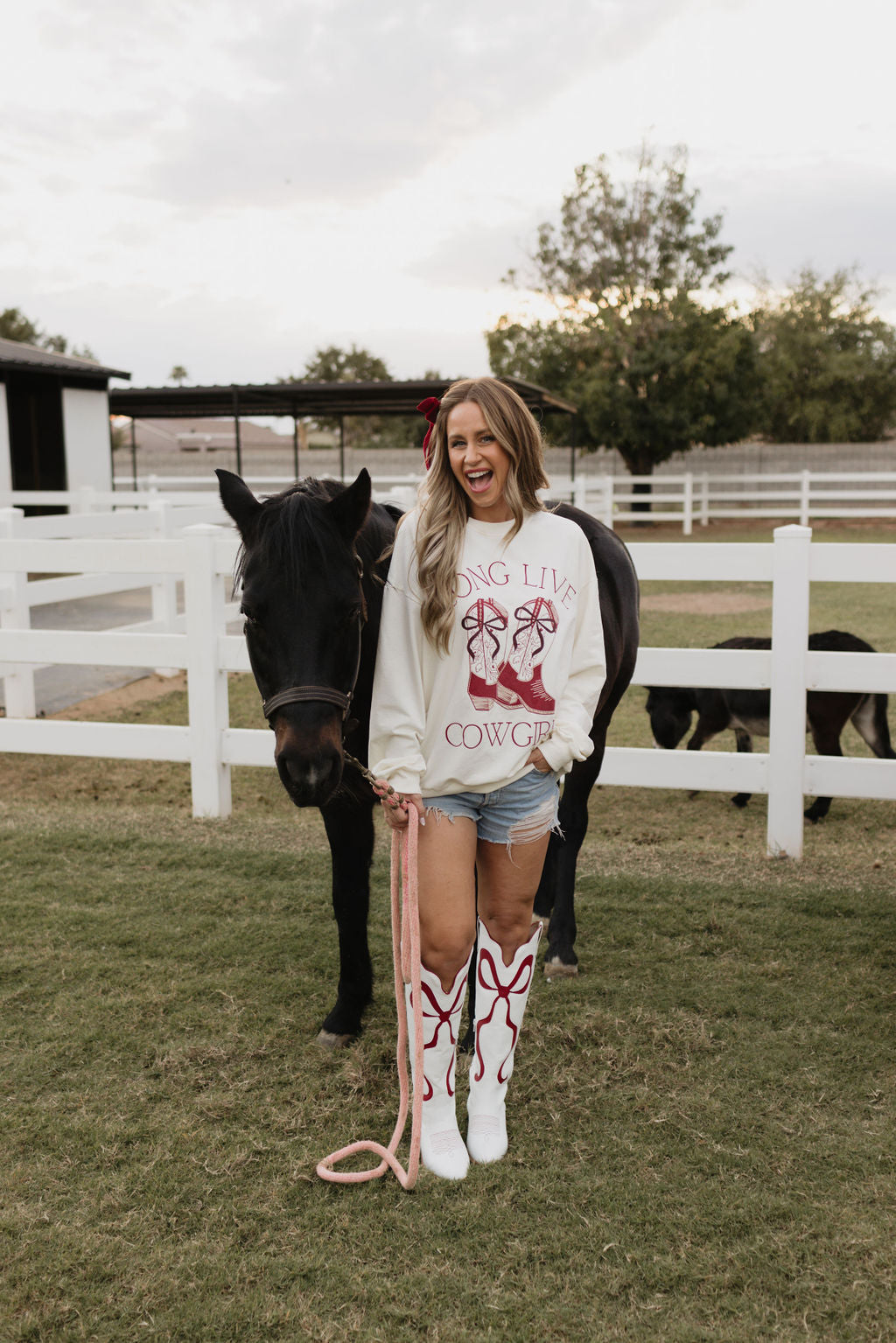 A woman in the Adult Crewneck by lolo webb, which features the phrase "Long Live Cowgirls," stands beside a black horse, holding its pink lead. She's wearing white boots and is laughing with her hair down in front of a white fence under a cloudy sky. There's also a matching children's version for young cowgirls.