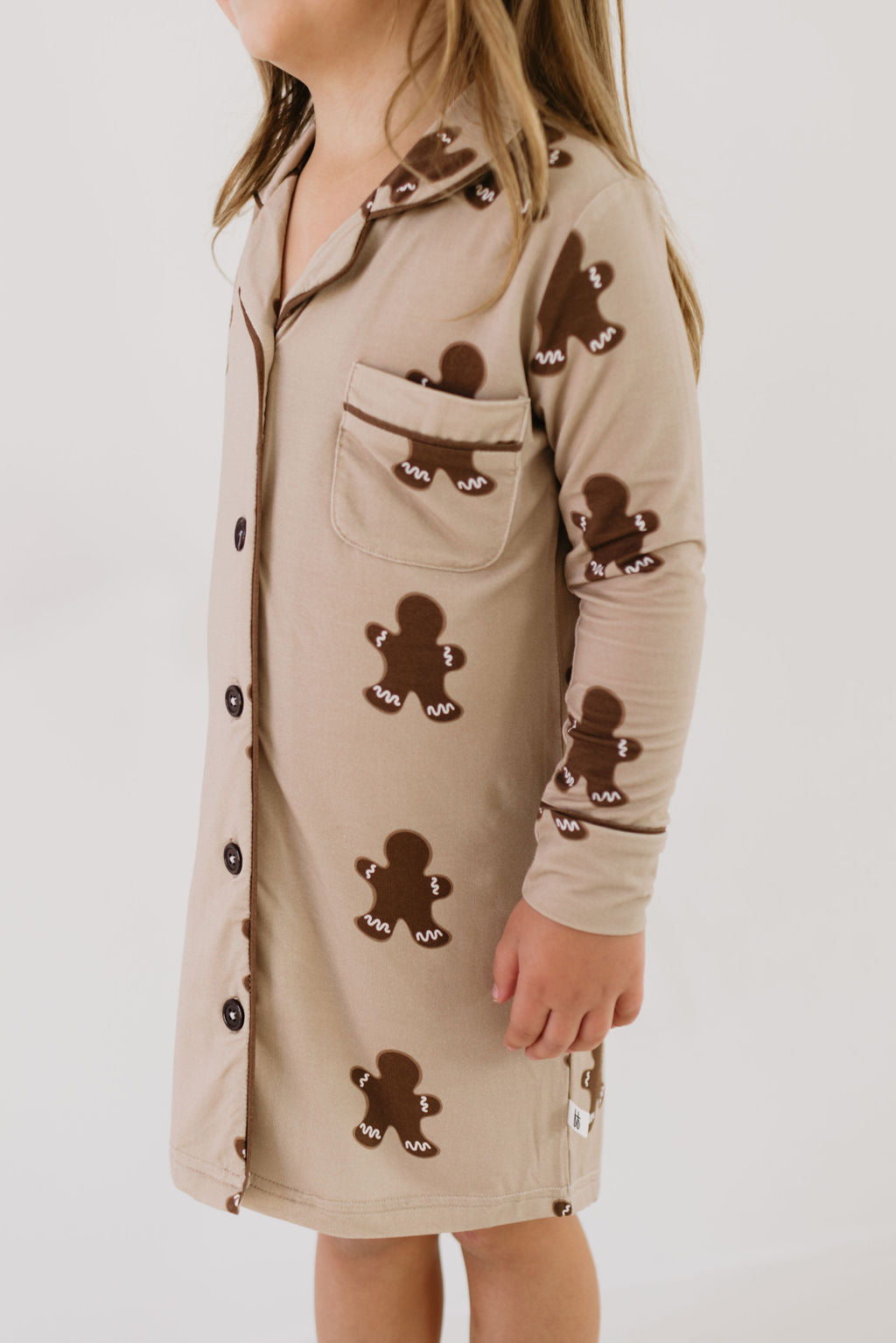 A young child wearing the Toddler Bamboo Sleeping Dress in a gingerbread pattern from forever french baby stands against a plain background. Made from soft bamboo fabric, this hypo-allergenic dress includes buttons down the front and a pocket on the chest. The child faces slightly to the side with comfort and ease.