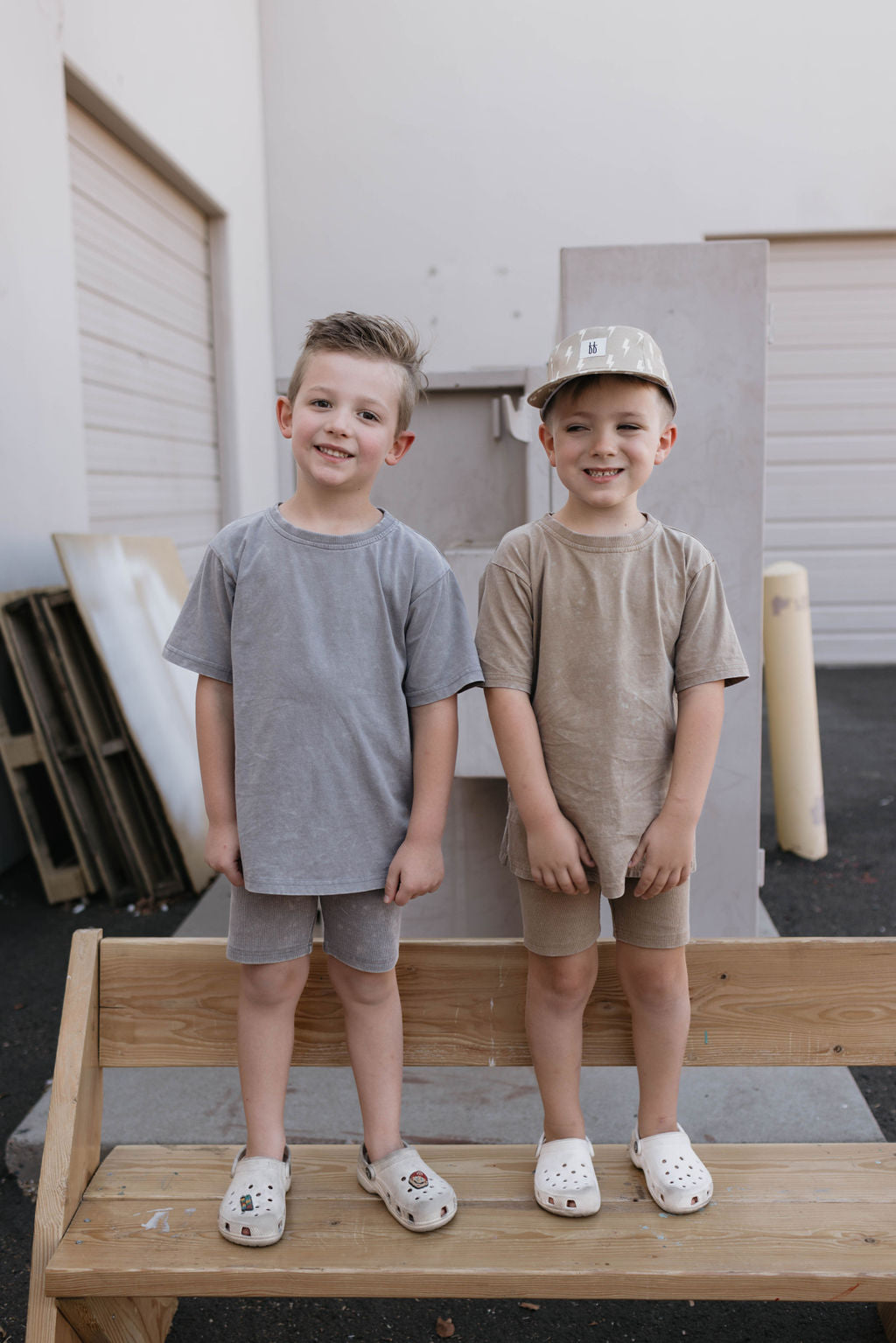 Two young boys stand on top of a wooden bench outdoors, both smiling and dressed in casual children's summer outfits. The boy on the left wears grey, while the boy on the right sports a *forever french baby* Children's Short Set in Vintage Washed Espresso with a matching cap. Both have white sandals on. A garage door is visible in the background.