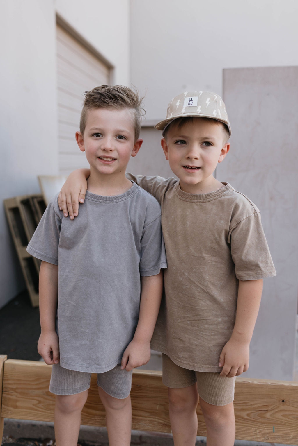 Two young boys stand next to each other outside, both smiling and wearing forever french baby's Children's Short Set in Vintage Washed Espresso. One boy has his arm around the other. The background features a building and some wooden panels, highlighting their charming summer outfits.