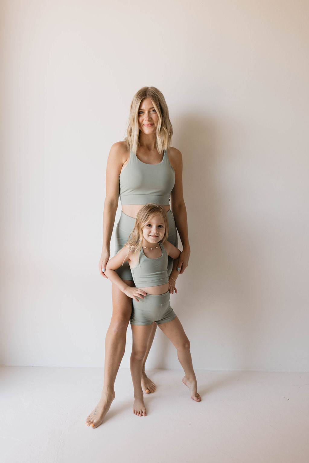 A woman and a young girl stand together against a plain white background, both dressed in matching Women's Workout Set | Green from forever french baby. The woman is positioned behind the girl, resting her arms on the girl's shoulders. Both are smiling, radiating happiness and unity in their coordinated active wear.