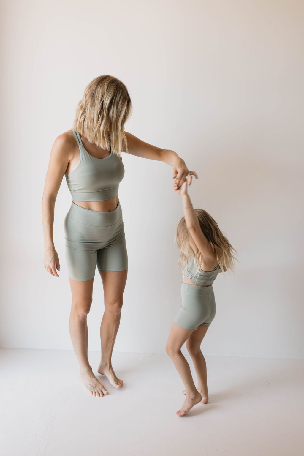 A blonde adult and a young girl, both sporting the Women's Workout Set in green from forever french baby, are inside a bright room with white walls. The adult holds the girl's hand as she spins around joyfully, creating a light-hearted atmosphere.