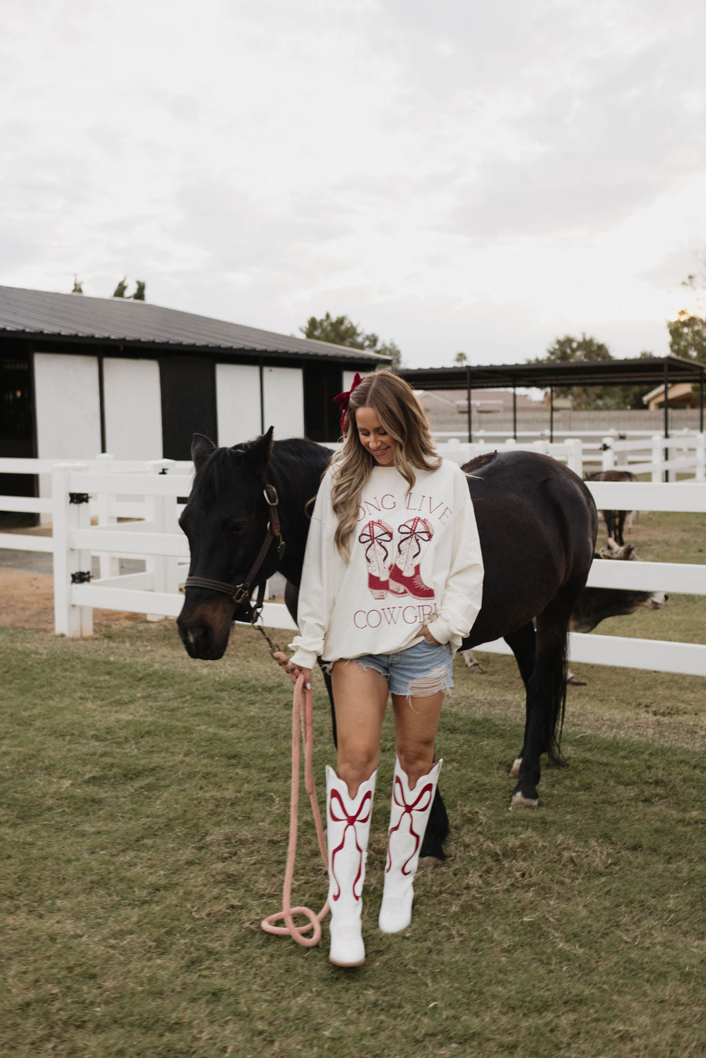 A woman wearing the "Long Live Cowgirls" adult crewneck by lolo webb, along with a matching children's version, stands next to a black horse. She is dressed in white boots with red designs and holds a pink lead rope on a grassy area near a white fence and barn.