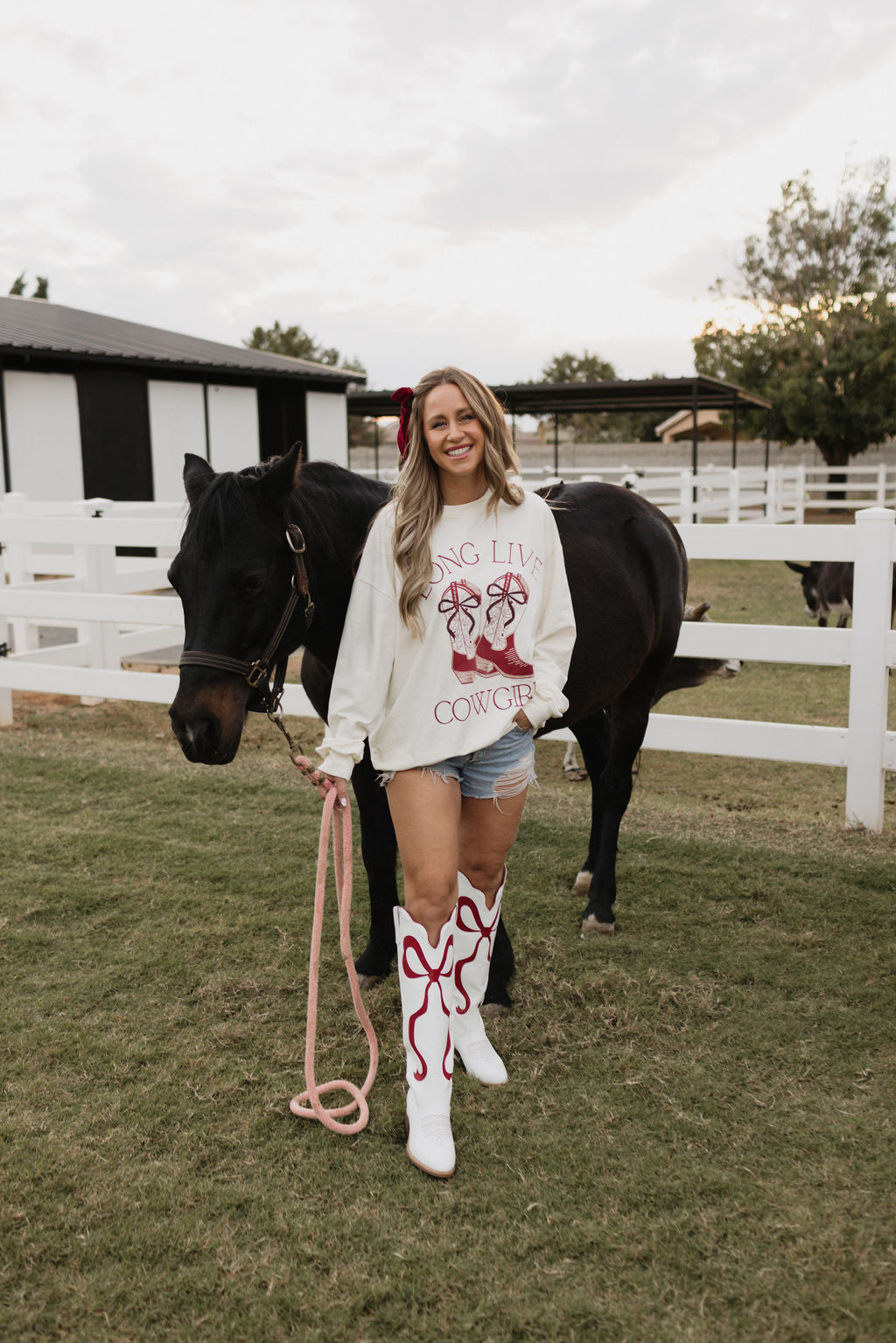 A woman stands beside a black horse in a grassy area, wearing the cozy Adult Crewneck from lolo webb, which proudly displays "Long Live Cowgirls." Her denim shorts and white cowboy boots with red designs add flair to her outfit. With a smile, she holds a pink rope, while a white fence and other animals complete the scene.