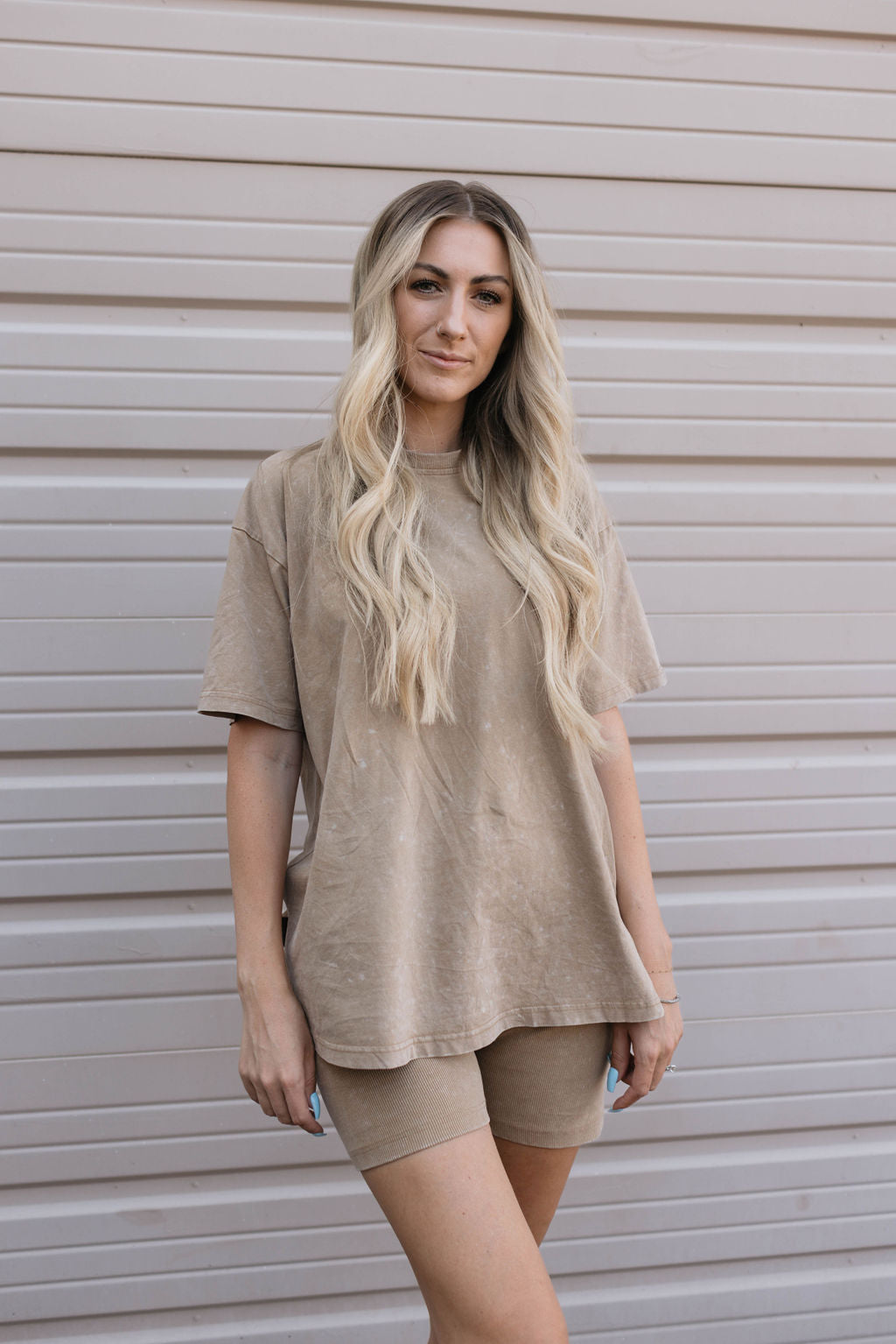 A woman with long, wavy blonde hair stands in front of a beige, metal garage door. She is wearing an oversized t-shirt and matching shorts from the Women's Short Set | Vintage Washed Espresso collection by forever french baby. She looks at the camera with a neutral expression and has her hands by her sides.