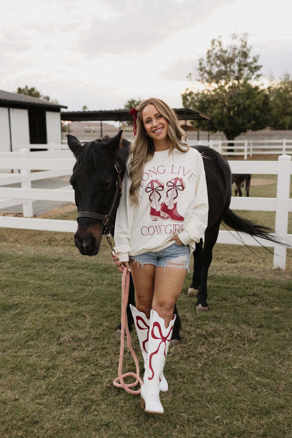 A woman smiles beside a horse on the farm, wearing the lolo webb Adult Crewneck featuring "Long Live Cowgirls" text. Her outfit is completed with denim shorts and white cowboy boots embellished with red designs. The backdrop includes a white fence and trees under a cloudy sky, suggesting there's a matching version for children nearby.