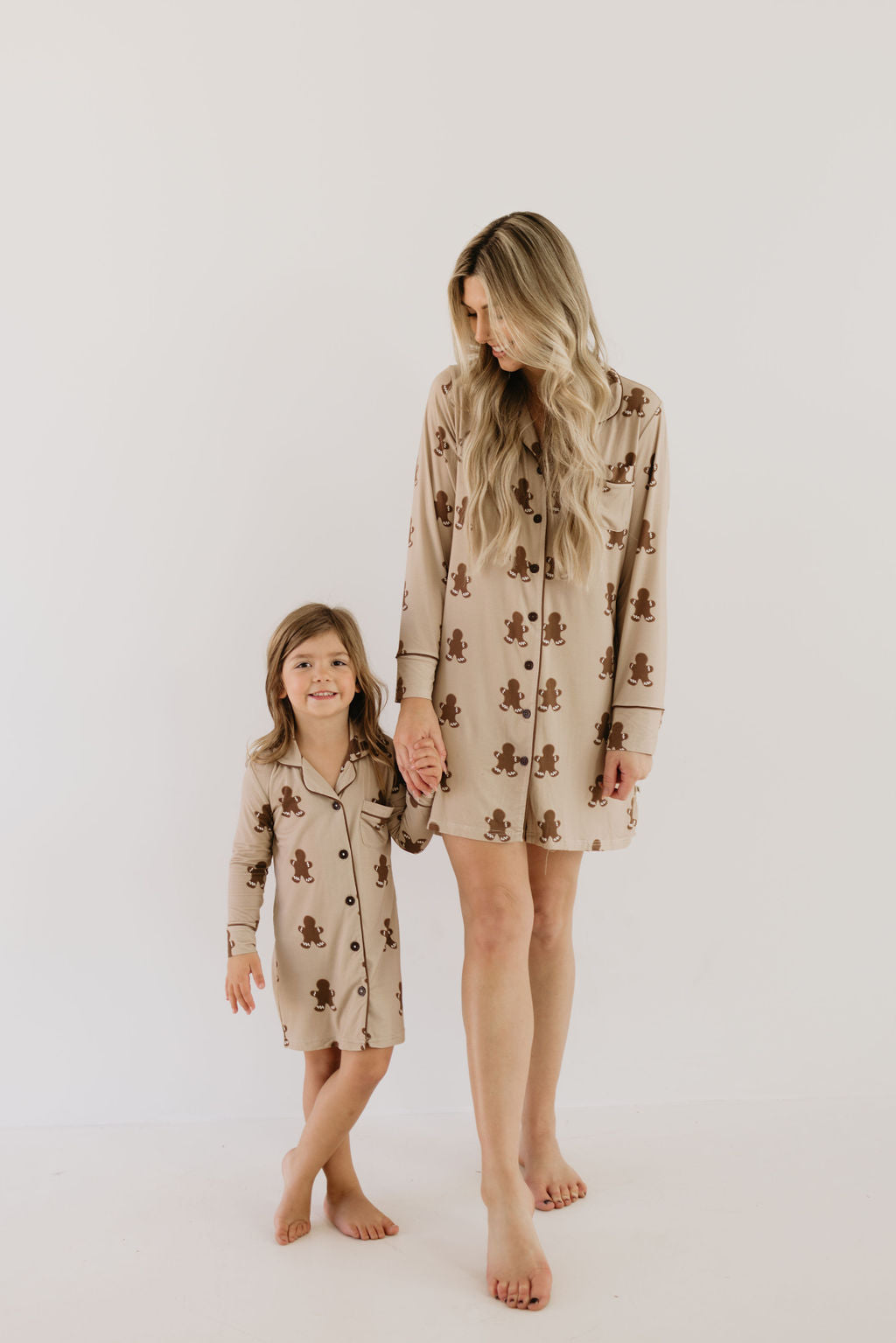 Against a plain white backdrop, a woman and a young girl with long hair stand barefoot. They are both wearing matching beige pajamas from forever french baby, specifically the Toddler Bamboo Sleeping Dress in the Gingerbread design. Made from hypo-allergenic bamboo fabric featuring gingerbread men, they hold hands while looking down and smiling.