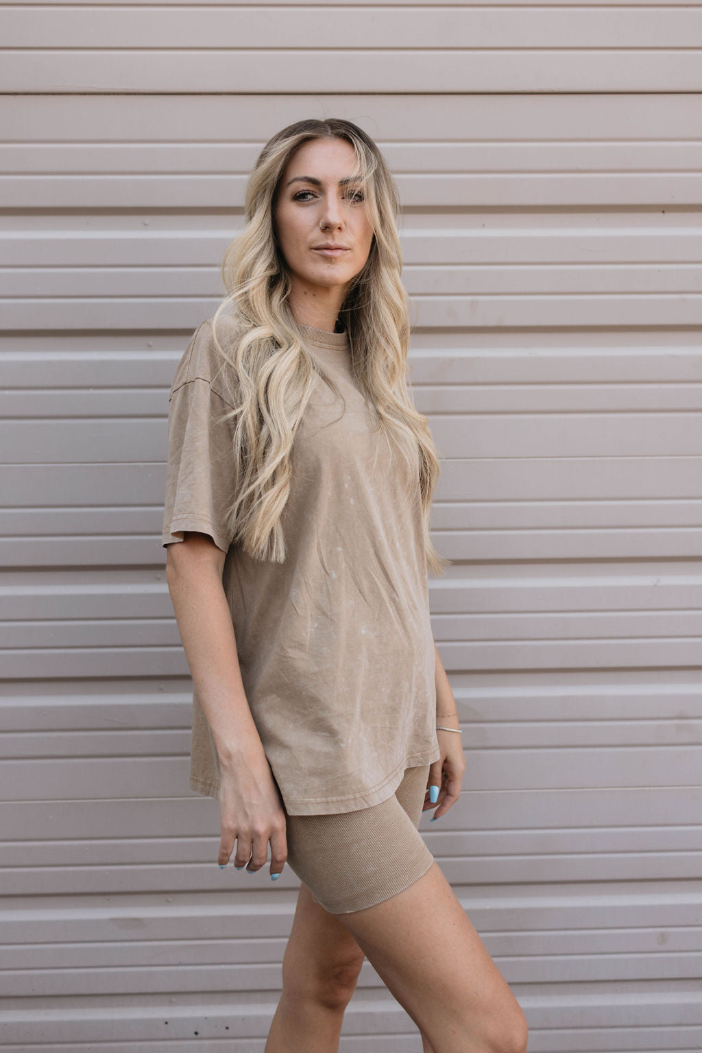 A person with long wavy blonde hair stands in front of a beige textured wall. Sporting the Women's Short Set in Vintage Washed Espresso from forever french baby, they exude effortless style. Their expression is neutral, casually posing as if embodying the timeless charm of Forever French Baby.