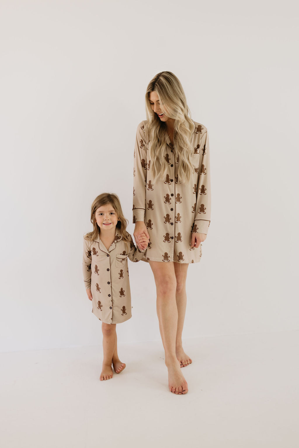 A woman and a young girl stand barefoot against a white background, both wearing matching hypoallergenic beige pajamas adorned with brown teddy bear patterns. The comfortable attire crafted from soft bamboo fabric, includes the "Toddler Bamboo Sleeping Dress | Gingerbread" by Forever French Baby. As the woman holds the girl's hand, they both smile warmly.