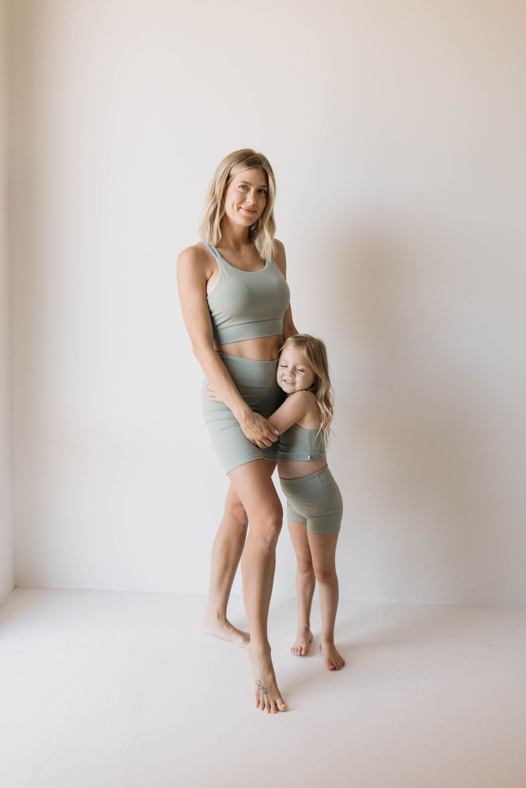 A woman and a young girl, both barefoot and clad in matching green activewear from the forever french baby's Women's Workout Set line, share a warm embrace while standing in a minimalist, white-walled room. The woman smiles gently while the girl grins broadly, looking up at her. The muted green tone of their outfits complements the serene ambiance.