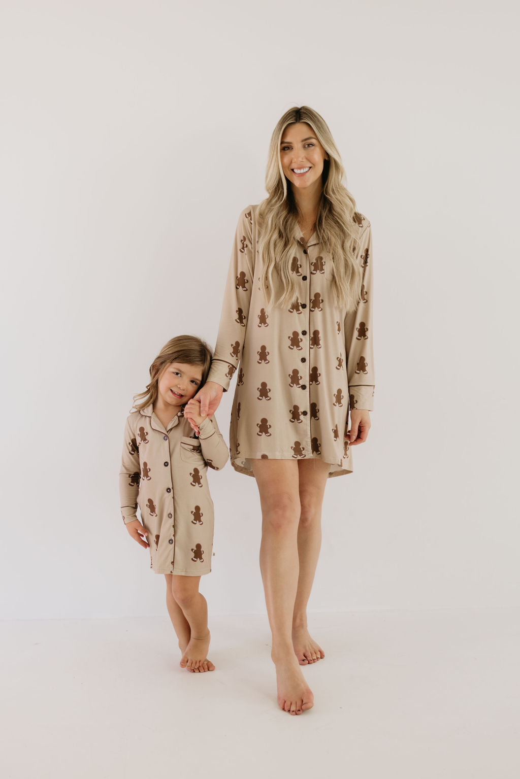 A woman and a young child, both barefoot, pose for the camera wearing matching gingerbread-patterned pajamas made from soft bamboo fabric. The toddler features the charming "Toddler Bamboo Sleeping Dress | Gingerbread" from forever french baby. The woman smiles while holding the child's hand, with both standing against a light-colored background that highlights their hypo-allergenic attire.