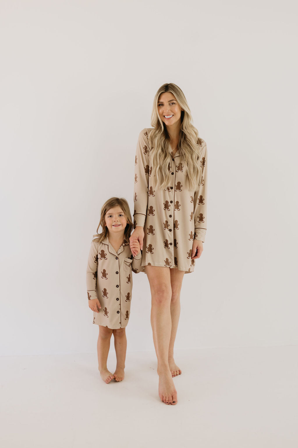 A woman and a child stand barefoot against a plain white background, both wearing matching long-sleeved pajama sets. The child's set is a Toddler Bamboo Sleeping Dress in the Gingerbread design from forever french baby. The pajamas feature a teddy bear pattern made from soft bamboo fabric. The woman has long blonde hair, and they hold hands.