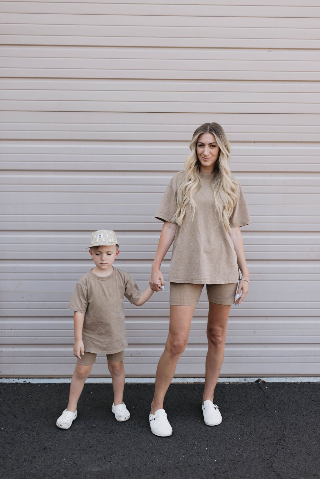 A woman with long blonde hair, wearing the Women's Short Set in Vintage Washed Espresso, stands holding hands with a young child. The child, dressed in the Forever French Baby collection in beige and a light-colored cap, is looking down. Both are standing in front of a striped beige garage door.