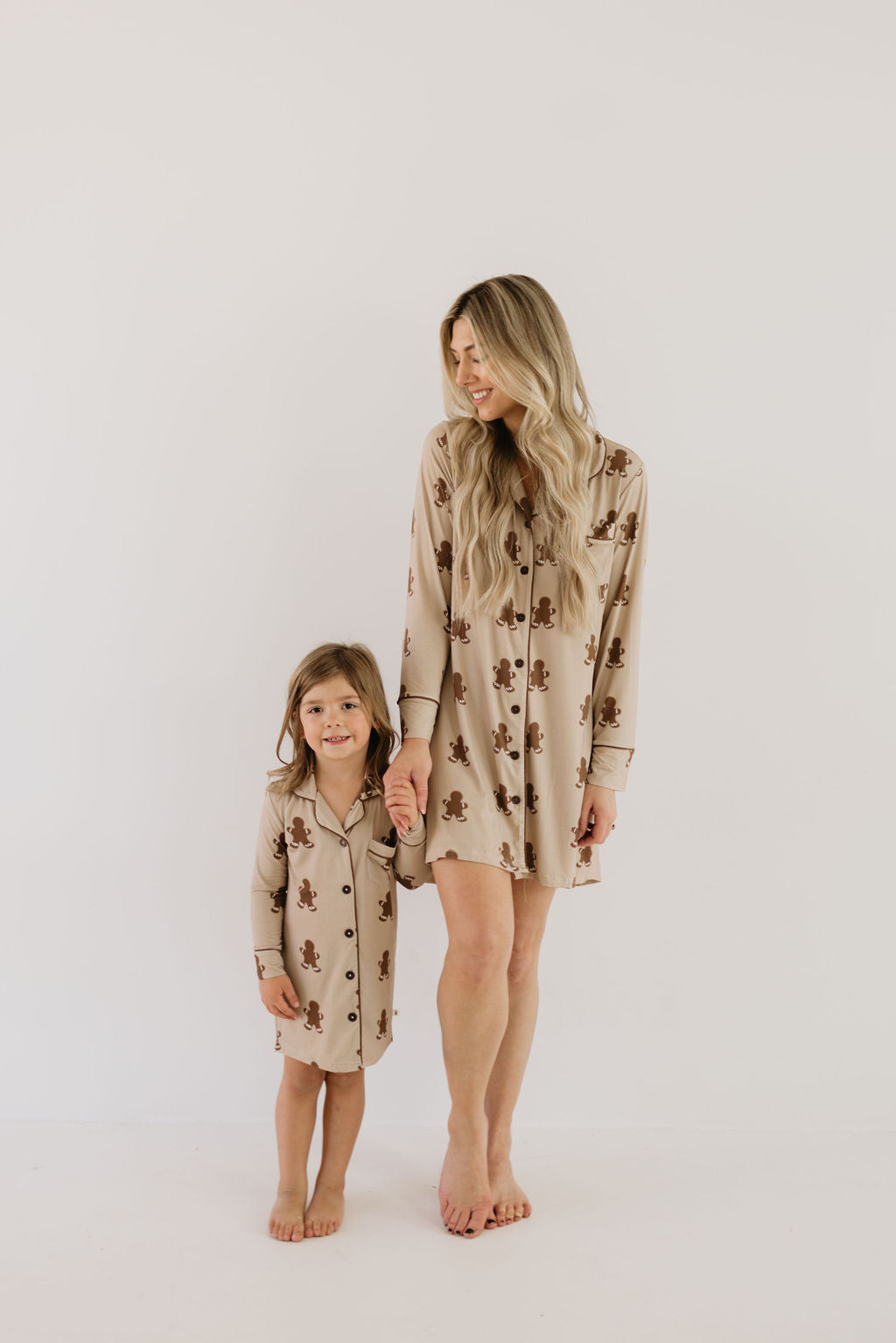 A woman and a young girl stand barefoot, holding hands and smiling in matching beige pajamas adorned with gingerbread patterns. These charming outfits, made by forever french baby from hypoallergenic bamboo fabric, guarantee comfort. The backdrop is a plain white wall.