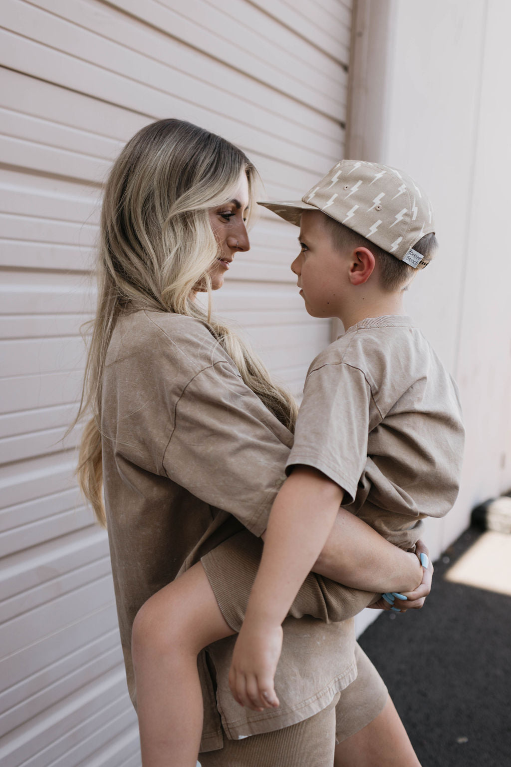 A woman with long blonde hair wearing the Women's Short Set in Vintage Washed Espresso from the Forever French Baby collection holds a young boy dressed in a tan outfit and cap. They are standing in front of a light-colored garage door, gazing at each other lovingly.