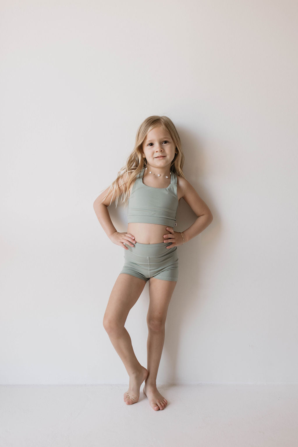 A young girl with long blonde hair stands against a plain white wall, wearing a light green Children's Workout Set from forever french baby. She is barefoot, with her right hand on her hip and her left arm relaxed by her side, looking confidently at the camera.