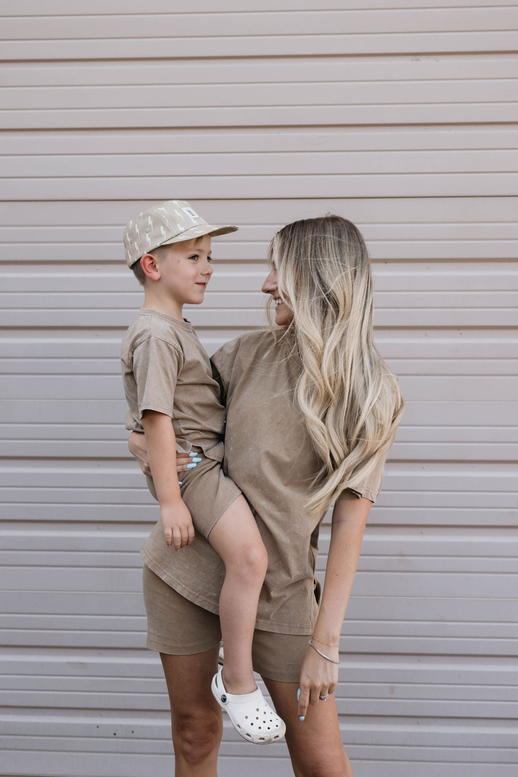 A woman with long blonde hair, dressed in a vintage washed espresso outfit from the Women's Short Set collection, is holding a young boy who is wearing a beige cap and matching outfit from Forever French Baby. Smiling at the boy, they stand in front of a light-colored striped wall. The boy sports crisp white shoes.