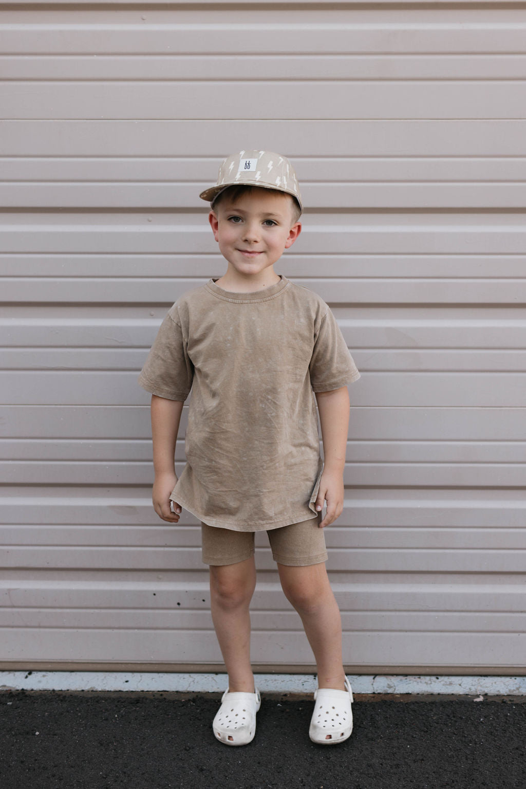 A young child stands in front of a beige garage door, donning a Forever French Baby Children's Short Set in vintage washed espresso, complete with white polka dots on the cap. The child pairs this adorable outfit with white clogs and smiles at the camera, hands at their sides, embodying the perfect children’s summer ensemble.