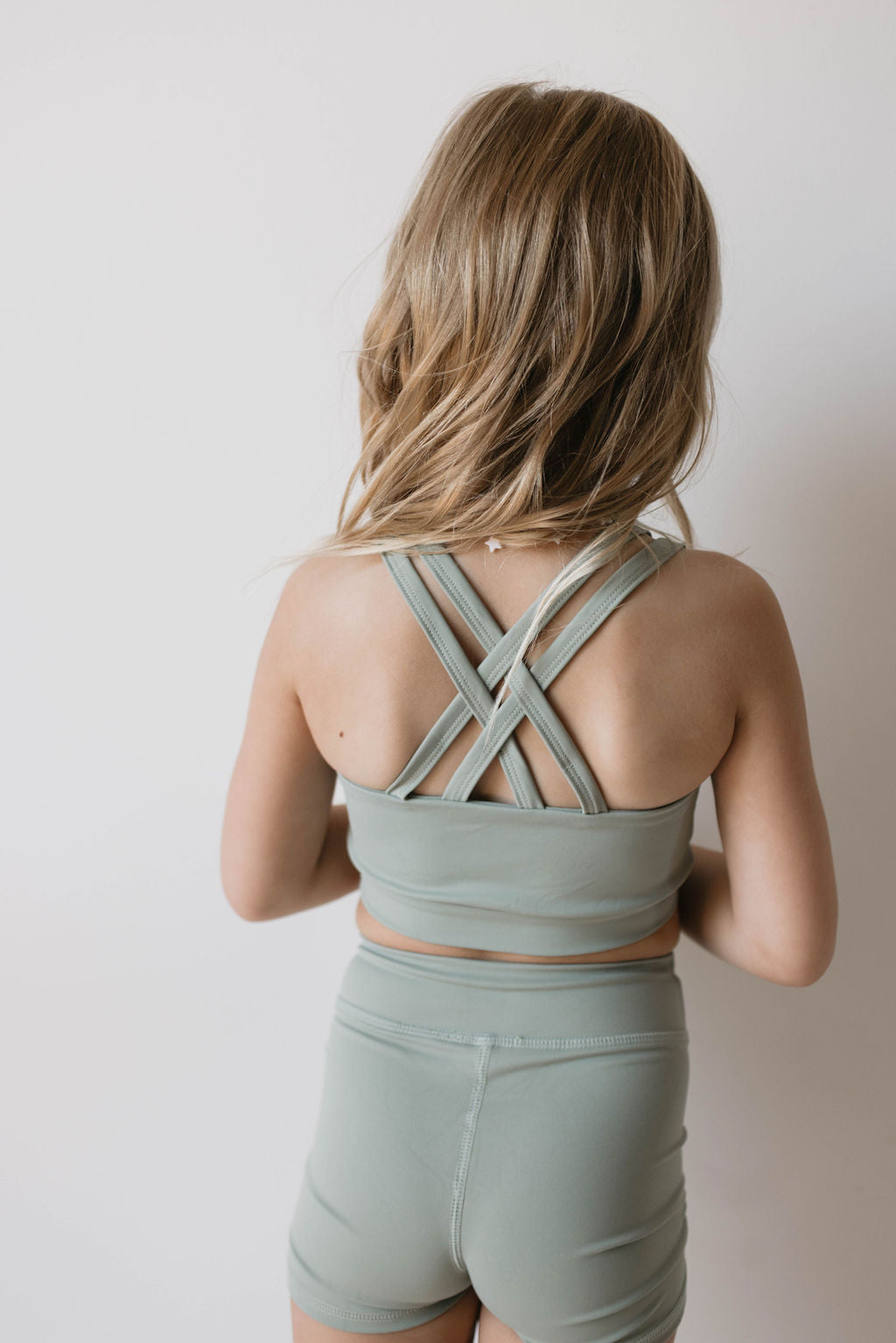 A young girl stands with her back to the camera, proudly showcasing the forever french baby Children's Workout Set | Green, consisting of a sports bra with crisscross straps and matching shorts. Her blonde hair is loose, cascading to her shoulders as she poses against a plain white background.