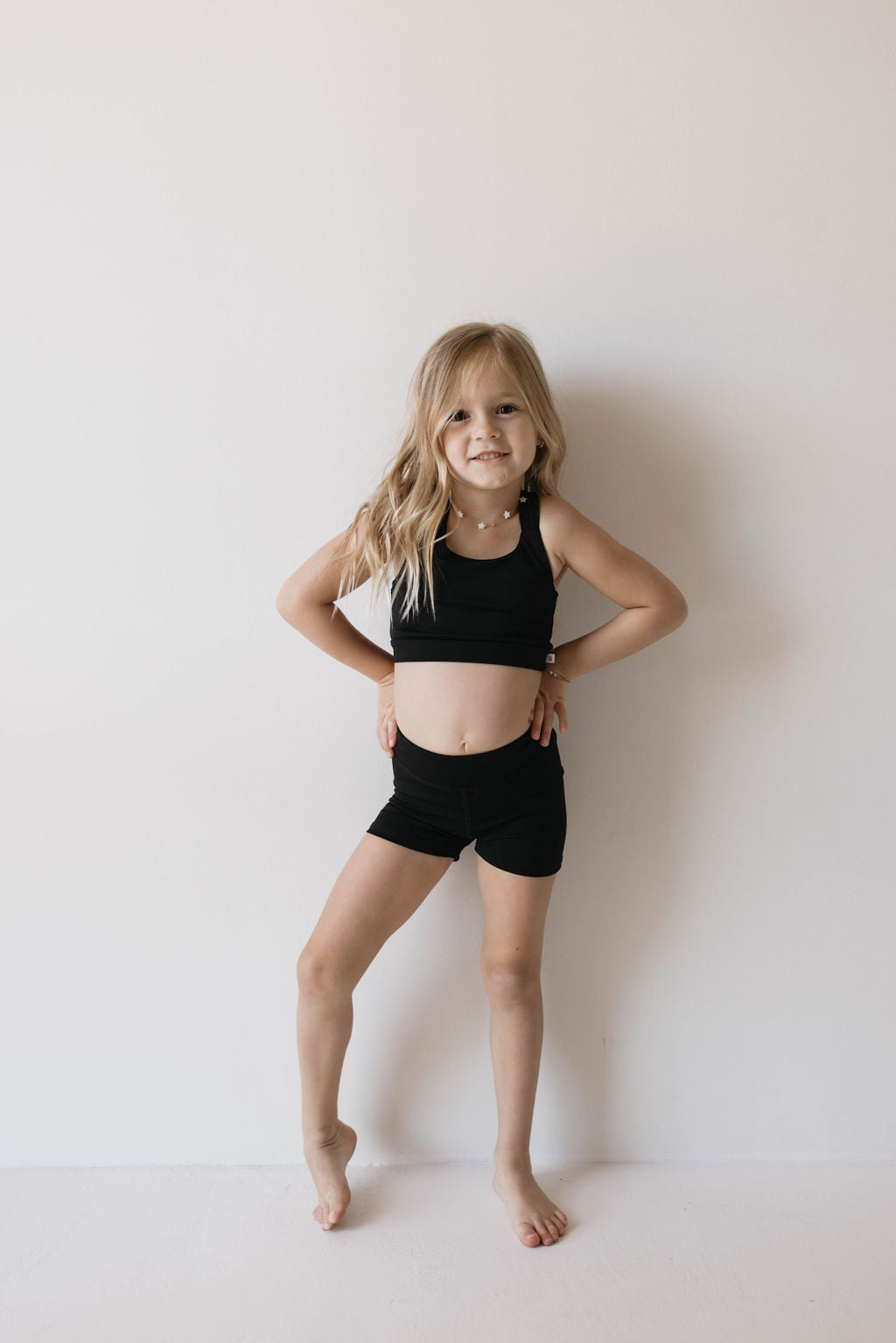 A young girl stands against a plain light-colored wall with her hands on her hips. She is wearing the Children's Workout Set in Black by forever French baby, which includes a crop top and matching shorts. She has long blonde hair and is barefoot, smiling slightly at the camera.