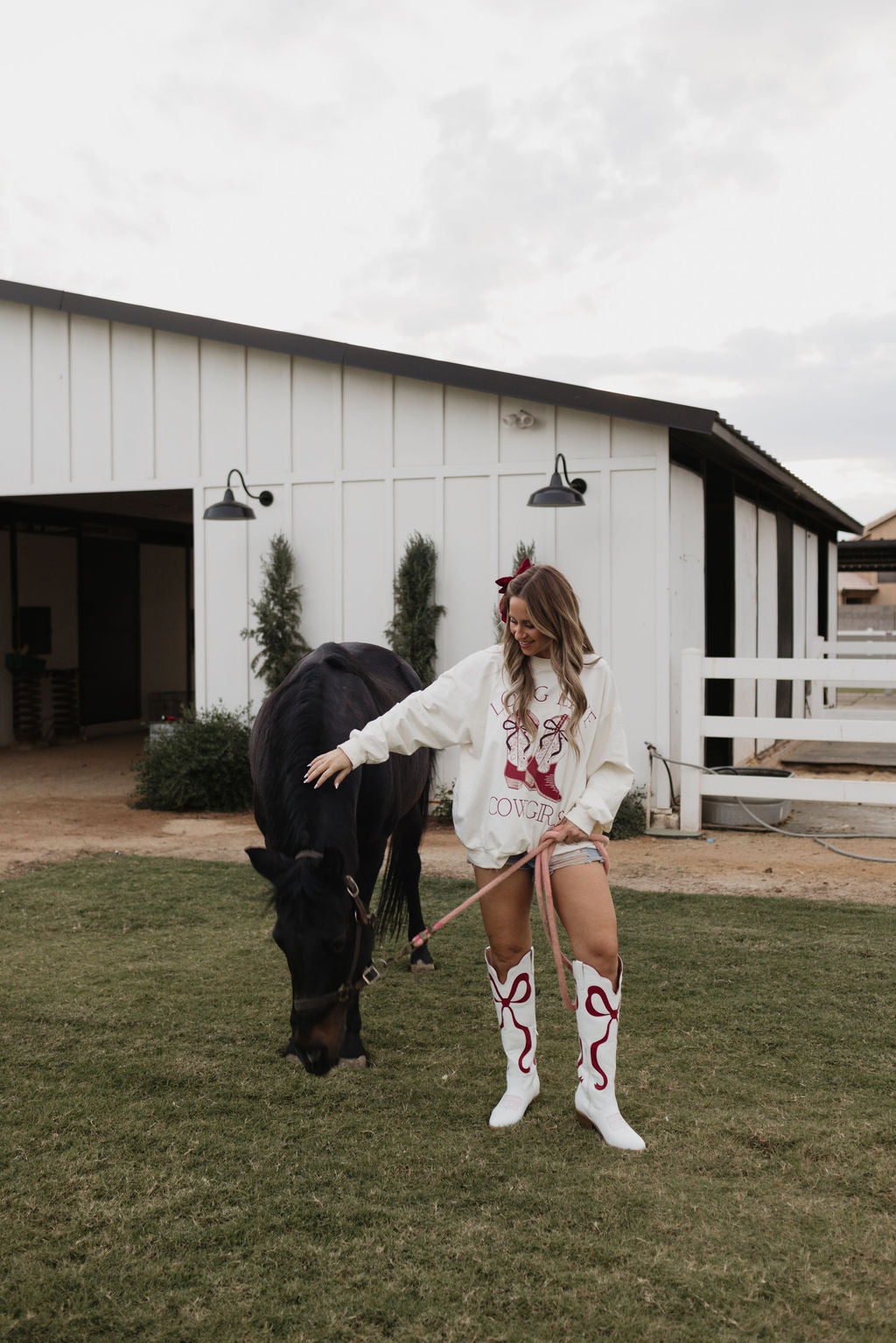 A woman wearing a pair of white cowgirl boots and the "Adult Crewneck | Long Live Cowgirls" sweatshirt by lolo webb stands on grass beside a black horse in front of a white barn. She holds the horse's reins and gazes down as it grazes. The sky is overcast, matching the mood of this tranquil moment.