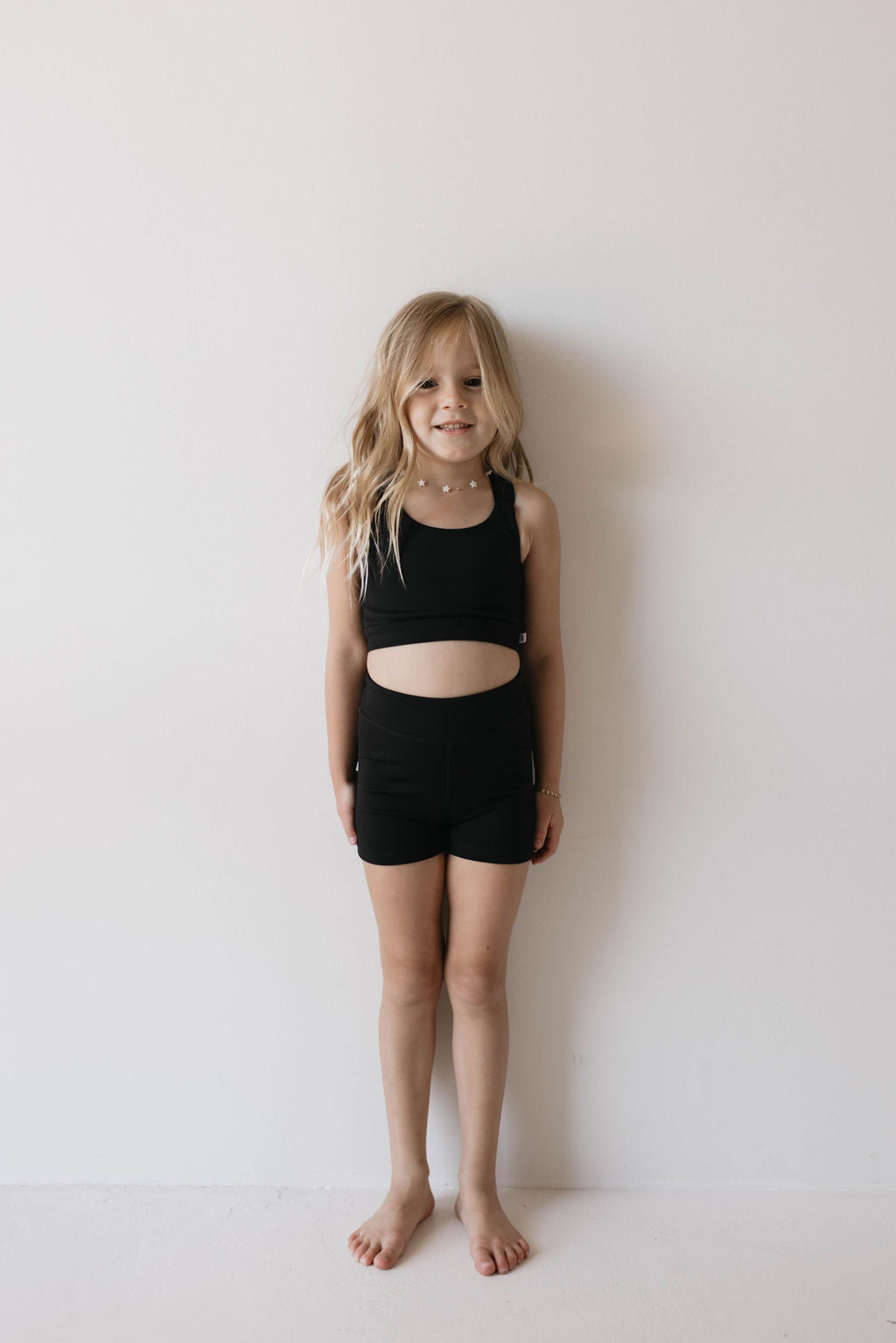 A young girl with long blonde hair stands barefoot against a plain white wall. She is smiling and wearing the Children's Workout Set in black, consisting of a sleeveless crop top and high-waisted shorts, from forever French baby.