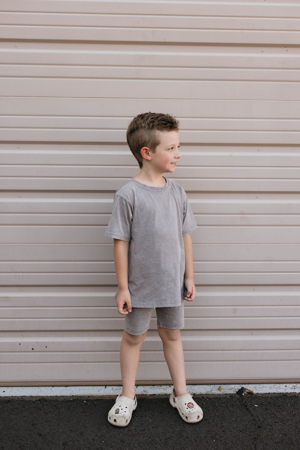 A young boy with short brown hair stands in front of a beige garage door, wearing a matching organic cotton grey t-shirt and shorts from the forever french baby Children's Short Set in Vintage Washed Steel. He is looking to his left and smiles slightly, also sporting white Crocs on his feet.