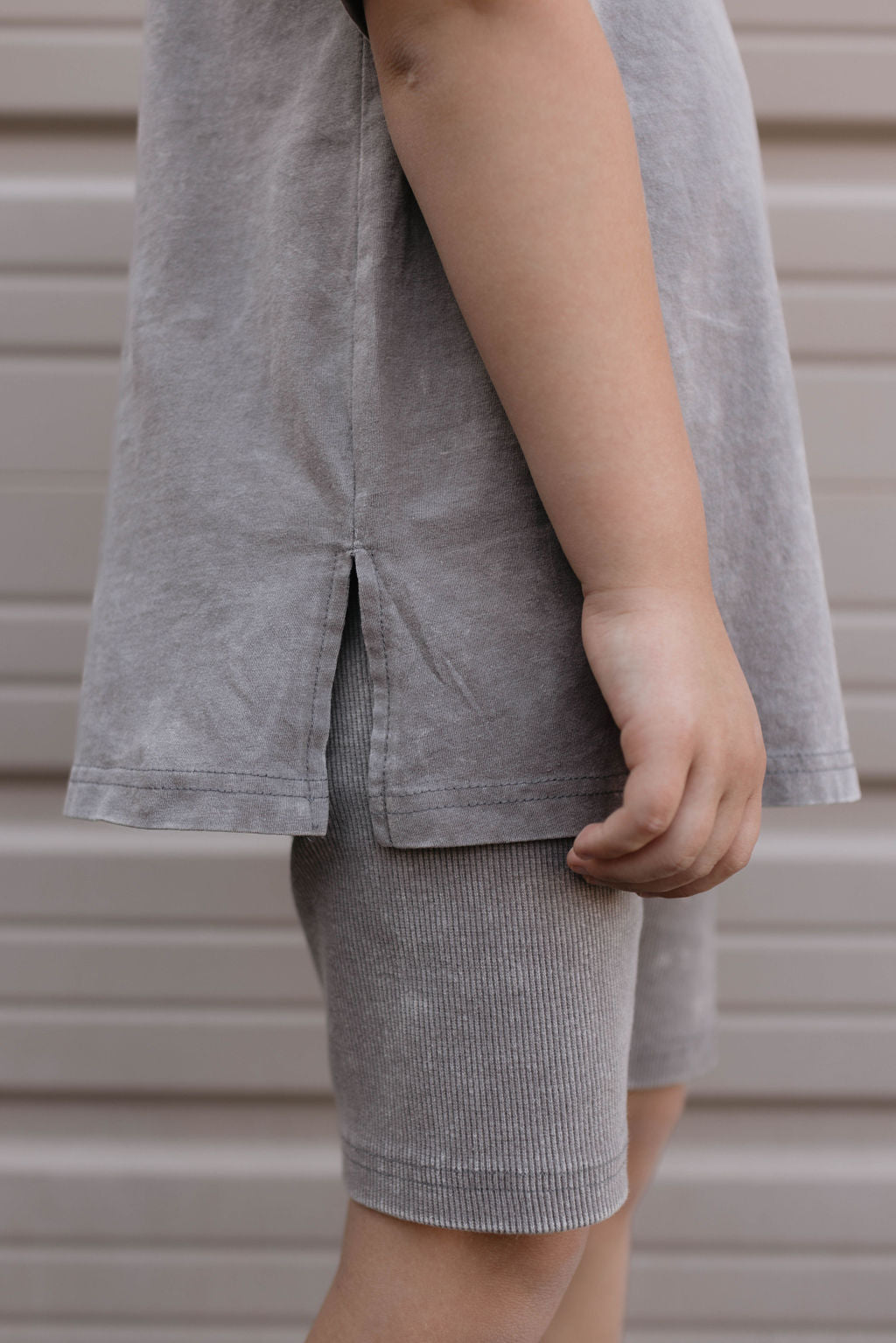 A close-up of a child's arm and side, wearing the Vintage Washed Steel T-shirt and ribbed grey shorts from the Children's Short Set by forever french baby. The background is a beige, paneled surface. Crafted from organic cotton, the T-shirt features a small slit on the side, with the child's hand relaxed and partially visible.