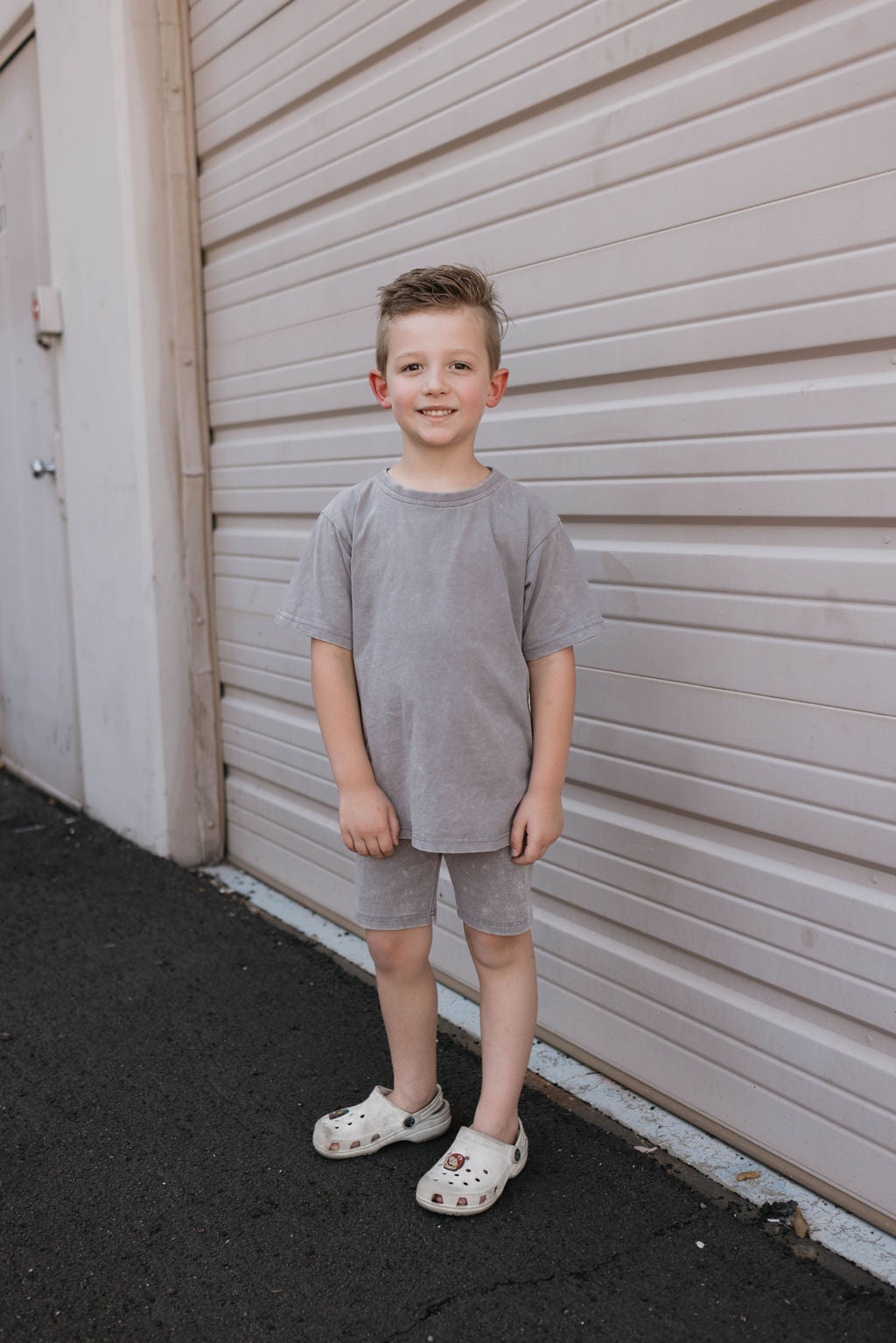 A young boy stands smiling in front of a large garage door, wearing a Vintage Washed Steel Children's Short Set by forever french baby, made from organic cotton. His hair is short and slightly tousled, and he completes his look with white slip-on shoes. The ground is paved, suggesting the setting is outdoors on a sunny day.