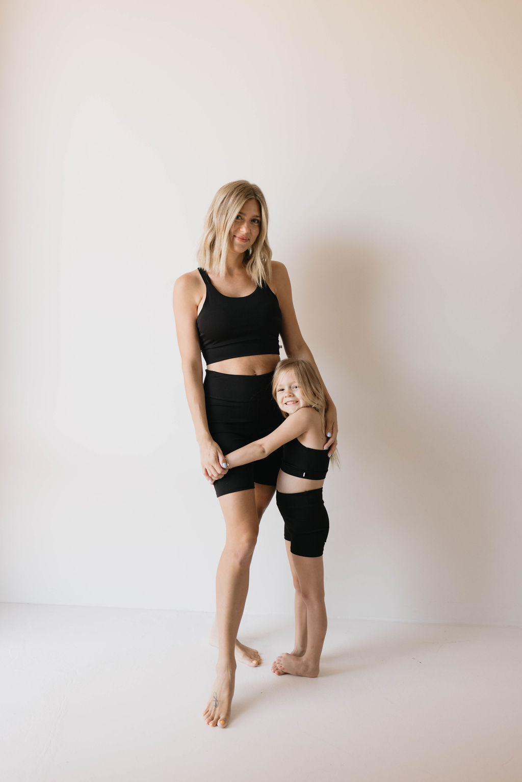 In a plain white background, a woman and a young girl dressed in matching black sports bras and shorts stand side by side. The woman wraps one arm around the girl’s waist while the girl hugs her closely. Both are barefoot and smiling at the camera, perfectly showcasing the Children’s Workout Set | Black from forever French baby – stylish kids active wear perfect for any activity.